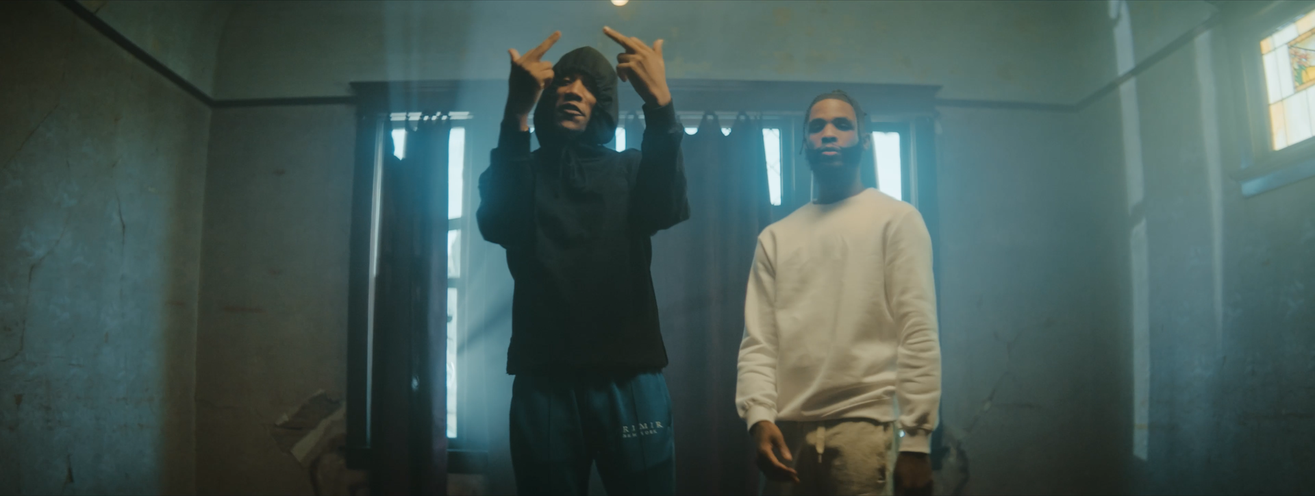 Two young men standing inside a worn, dimly-lit room with peeling walls and a window with sunlight streaming through. One is wearing a black hoodie with the hood up, making gestures with his hands towards his head. The other is wearing a white sweatshirt and has a beard, standing with one hand partially visible.
