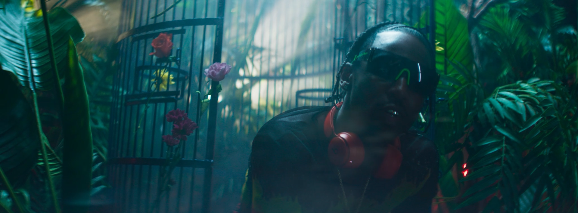 A person wearing sunglasses and headphones sitting amidst lush green tropical plants and flowers, with a background of a cage or decorative structure.