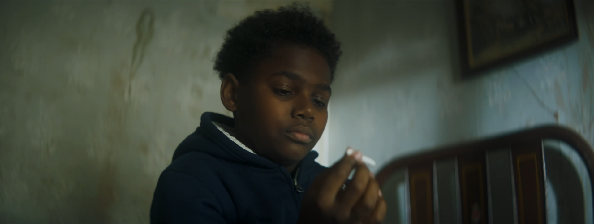 A young boy with curly hair and dark skin looks at an object in his hand with a serious expression in a room with a greenish wall.