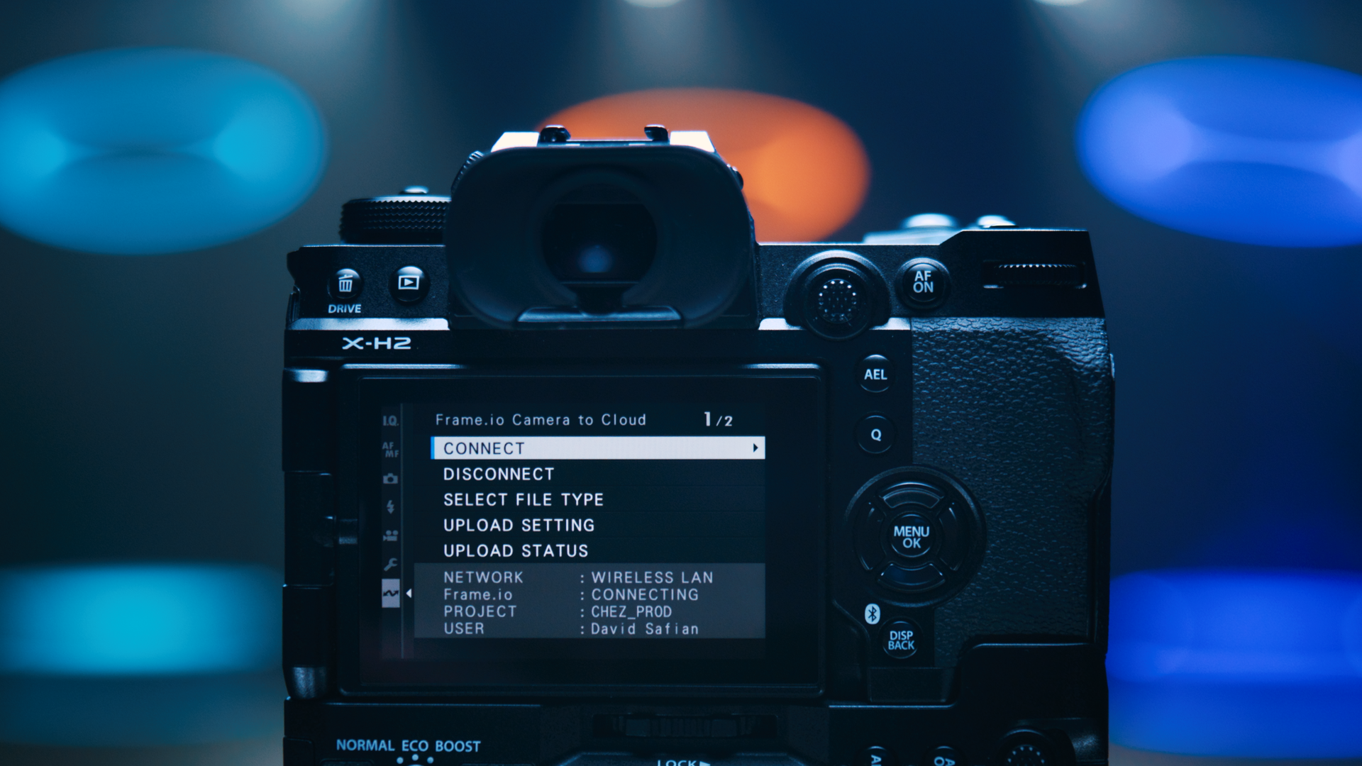 A digital camera showing a menu on its screen with options to connect, disconnect, and upload files, against a blurred background of colorful lights.