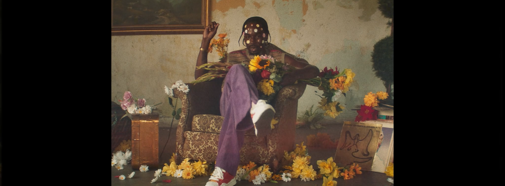 Person sitting on an ornate armchair surrounded by flowers, holding a bouquet, with a wall featuring a framed painting behind them.