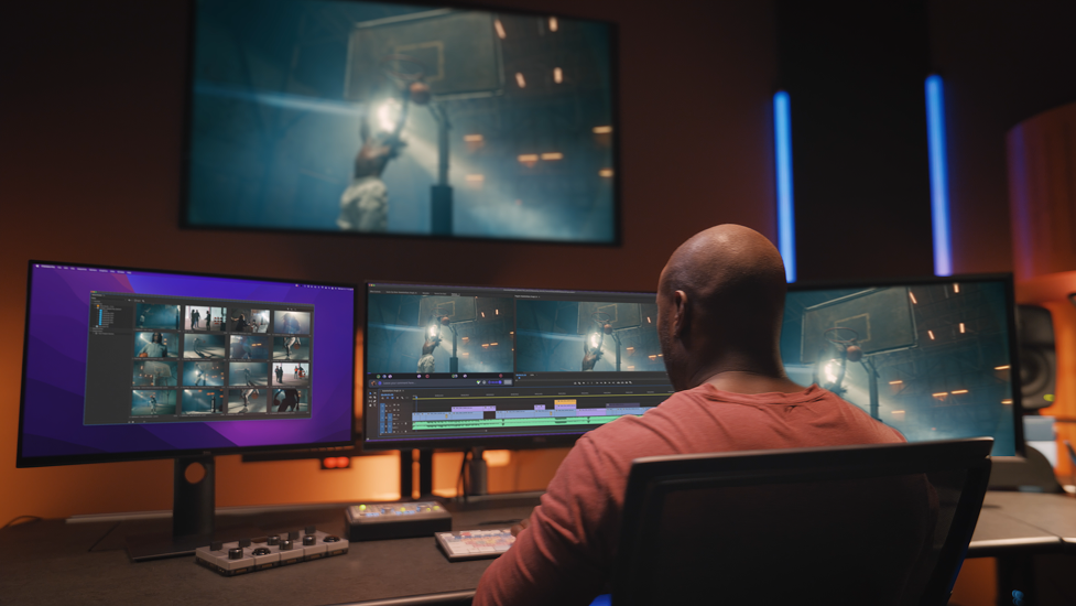 A person working on video editing with three computer monitors displaying a basketball game and editing software, in a dimly lit room with a large screen showing the same game in the background.