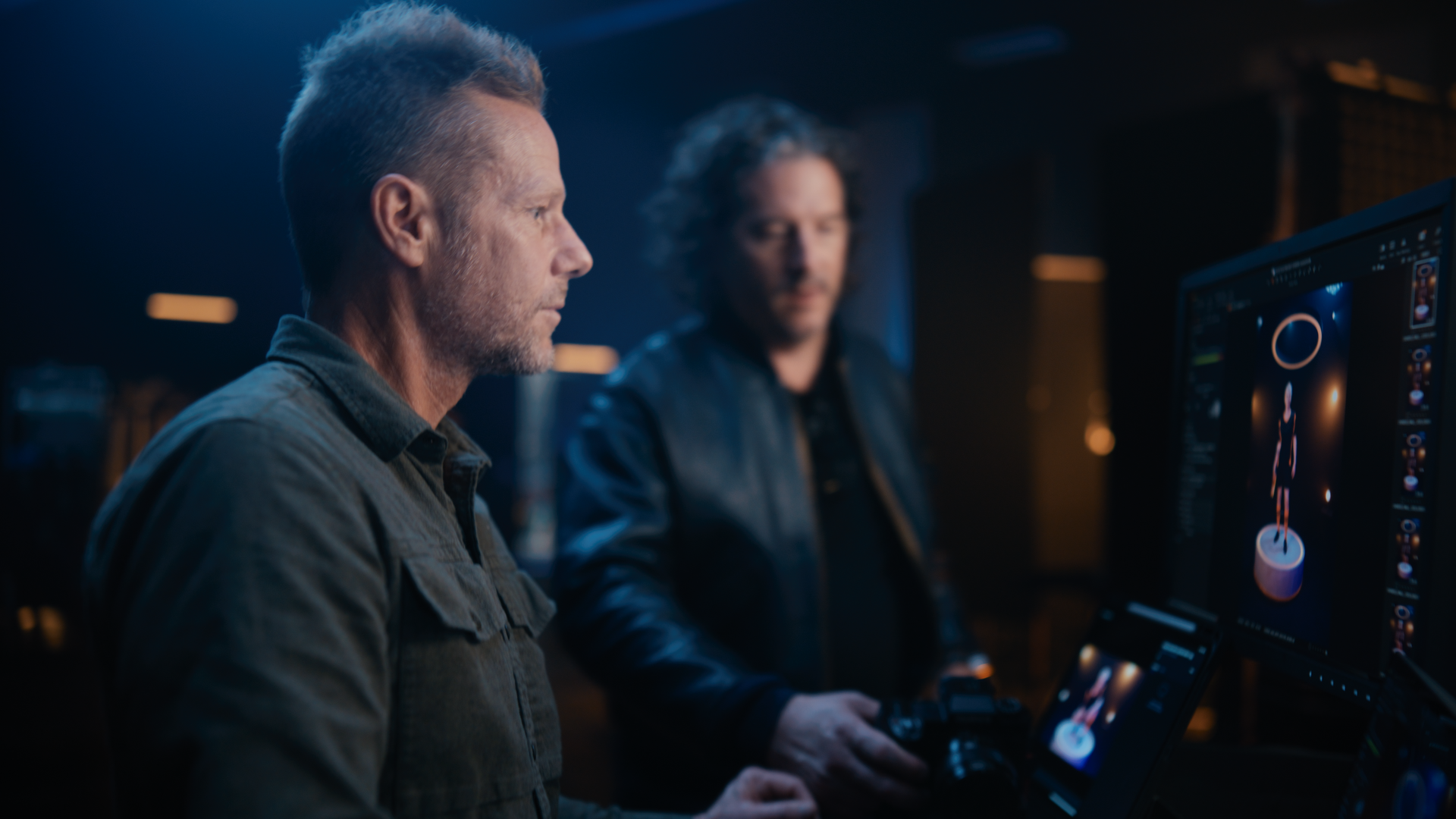 Two men working on a computer with a digital audio workstation on the screen in a dark studio.