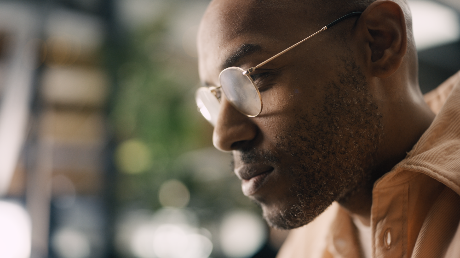 Close-up profile of a man with glasses and a beard, wearing a light brown jacket, with a blurred outdoor background.