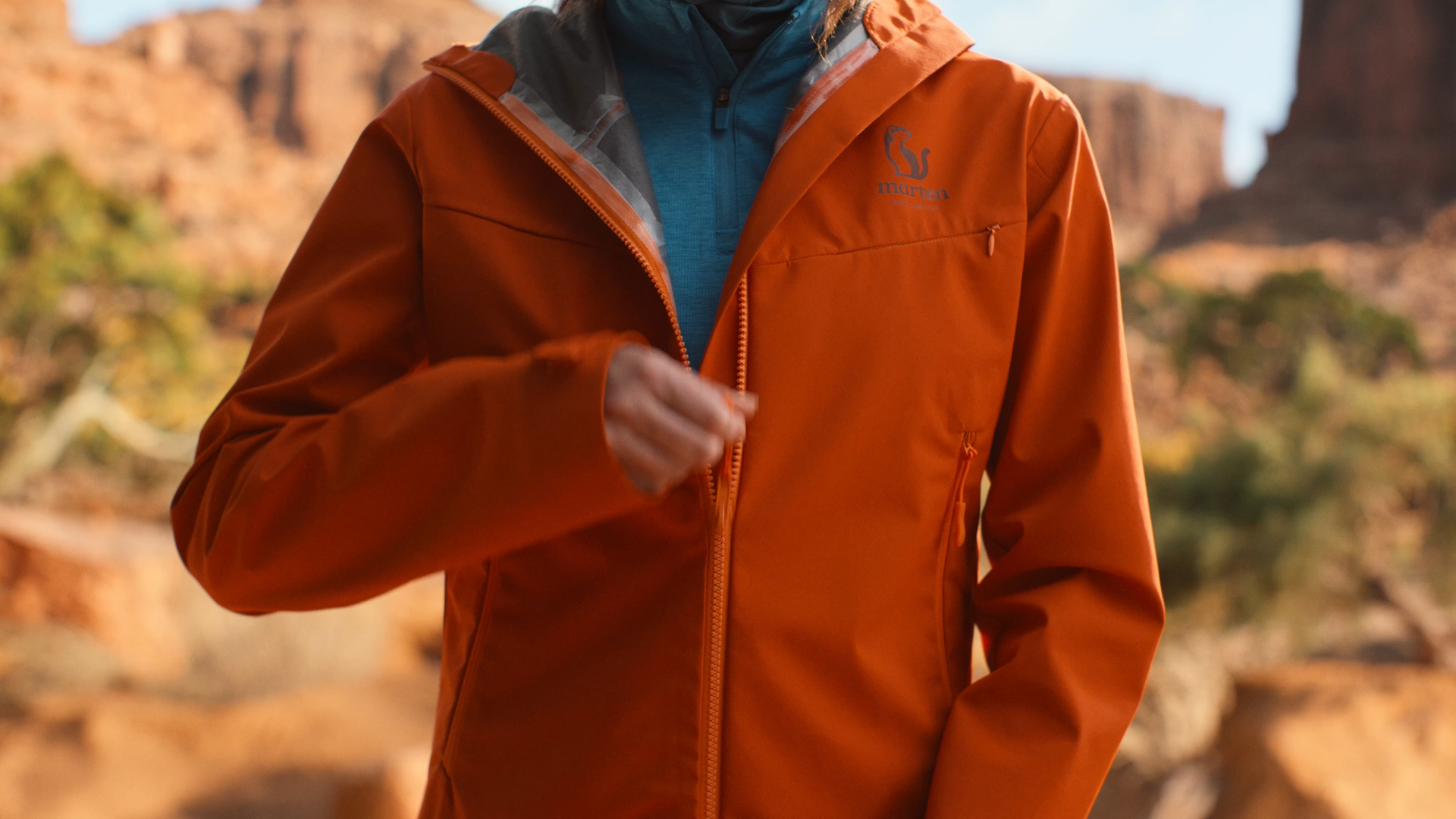 A person wearing an orange outdoor jacket with a gray hood and a blue shirt underneath, standing outdoors with rock formations in the background.