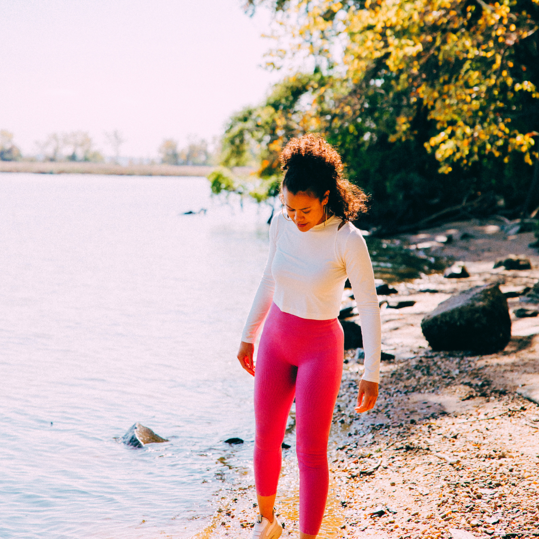 A woman in pink leggings and a white long-sleeve shirt walking on a rocky shoreline beside a lake with trees in autumn colors.