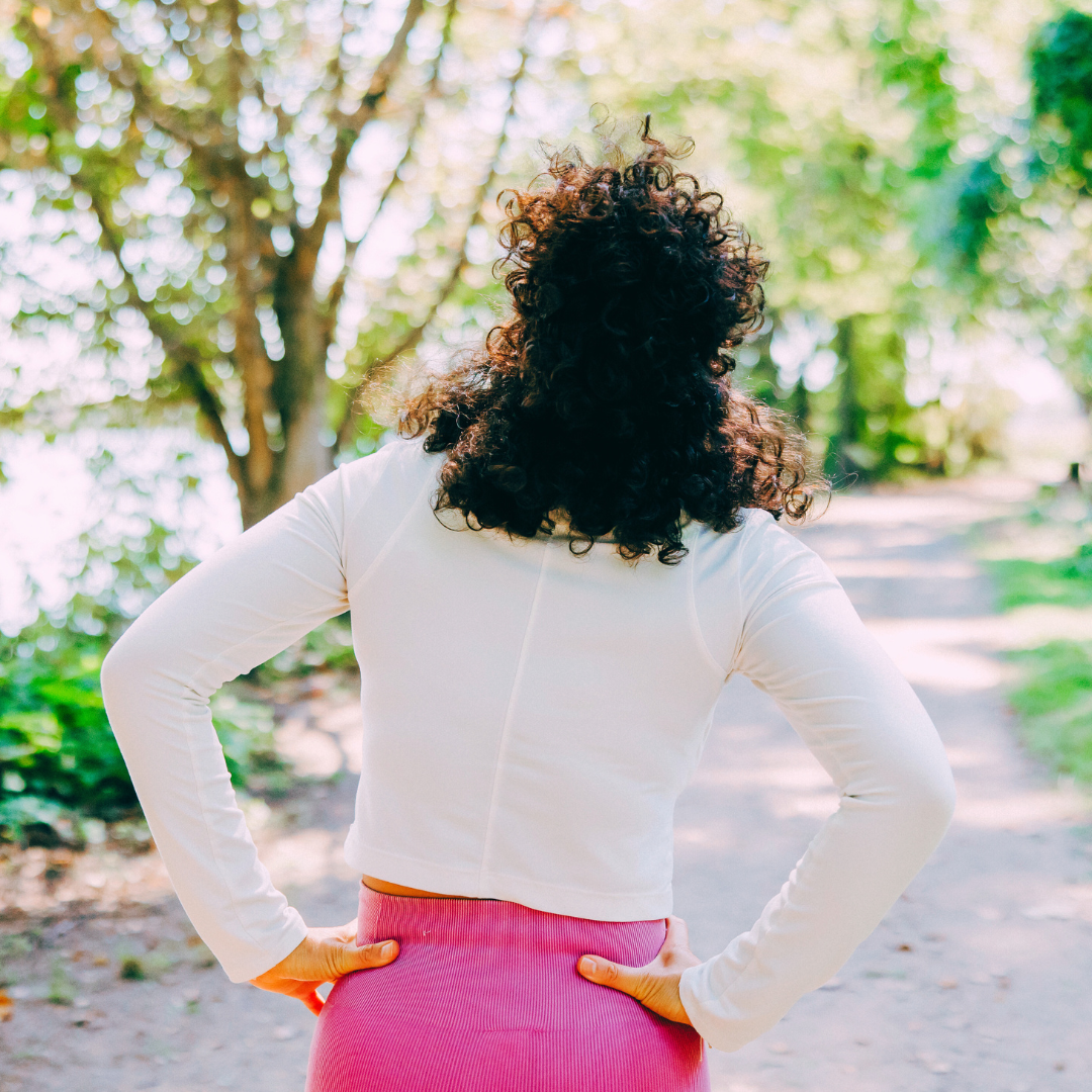 A woman with curly dark hair and a white long-sleeve top standing with her hands on her hips in a park with trees and sunlight.