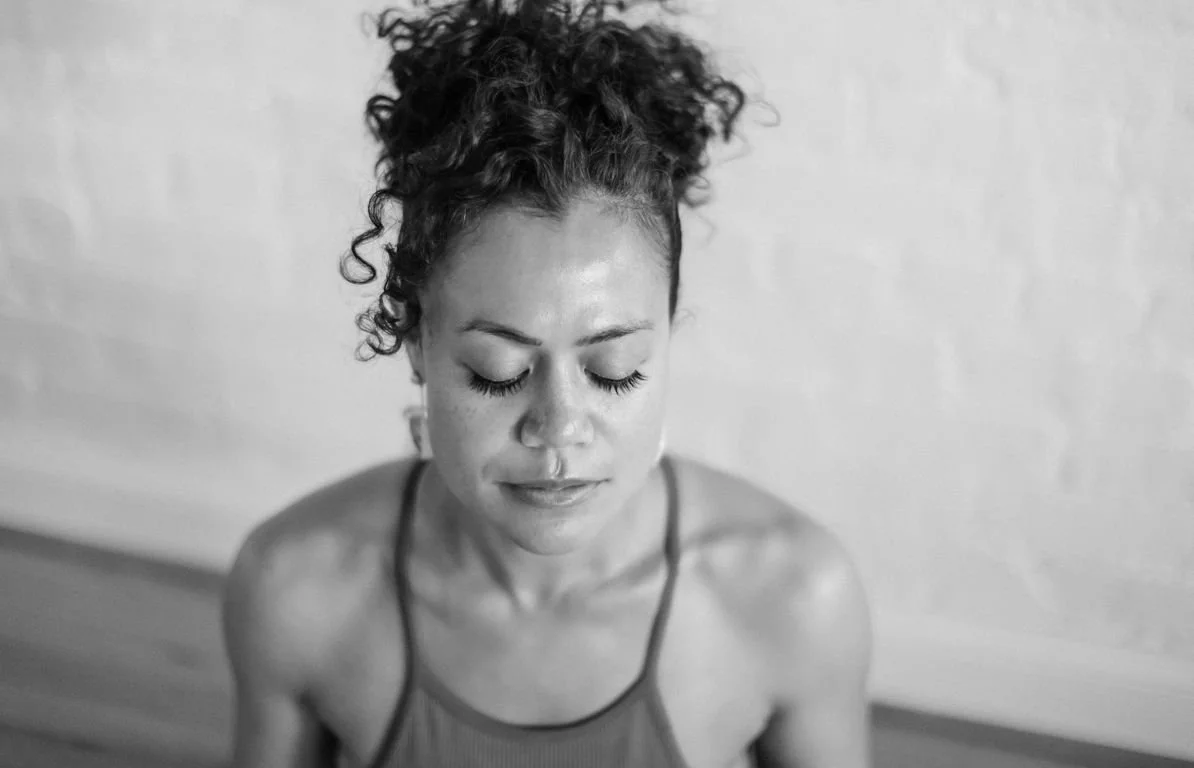 A black and white photo of a young woman with curly hair and closed eyes, wearing a tank top, standing against a plain wall with a peaceful expression.