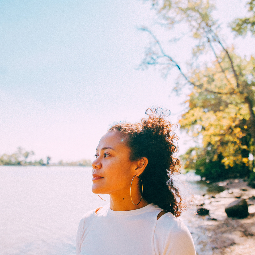 A woman with curly hair and large hoop earrings stands by a body of water on a sunny day, with trees in the background.