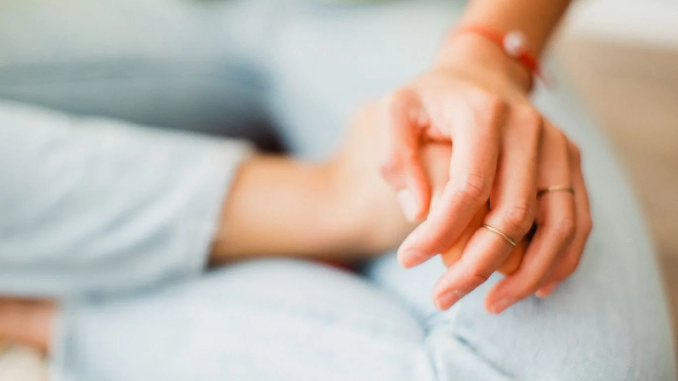 Close-up of a person’s hand resting on their knee, with visible rings and an orange bracelet.
