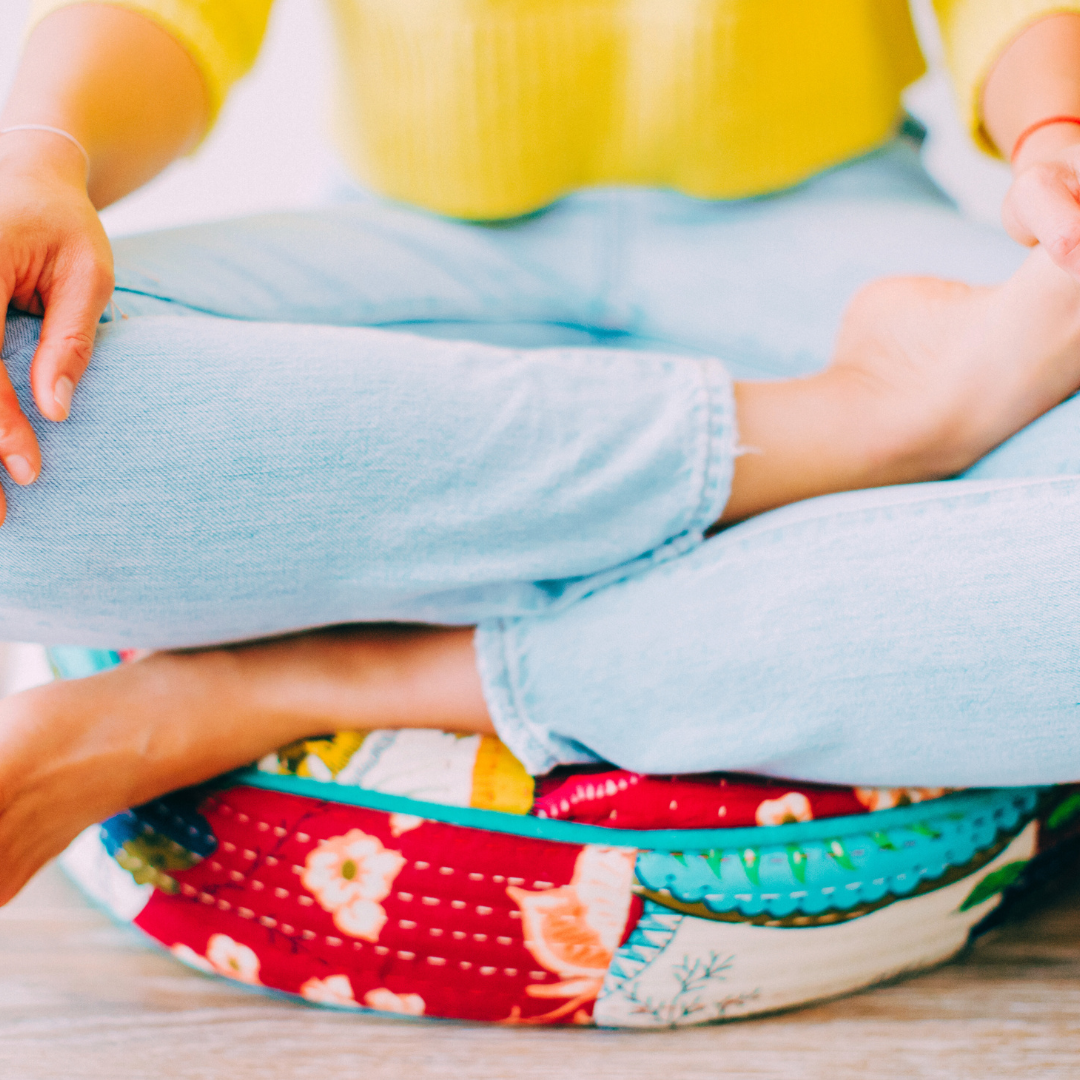 Close-up of a person practicing meditation or yoga, seated cross-legged on a colorful fabric cushion, wearing light blue jeans and a yellow top.