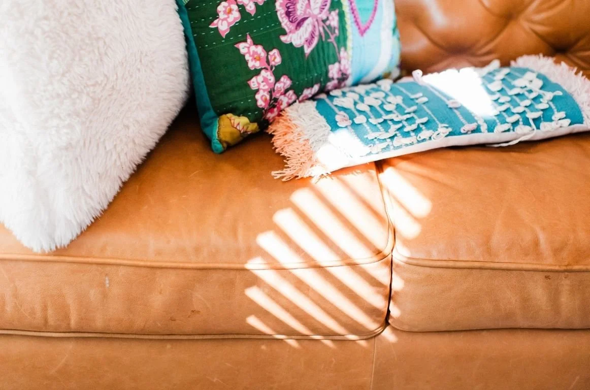 A tan leather couch with colorful pillows, including a green pillow with pink flowers and a blue pillow with white fringes, with sunlight creating linear shadow patterns on the couch.