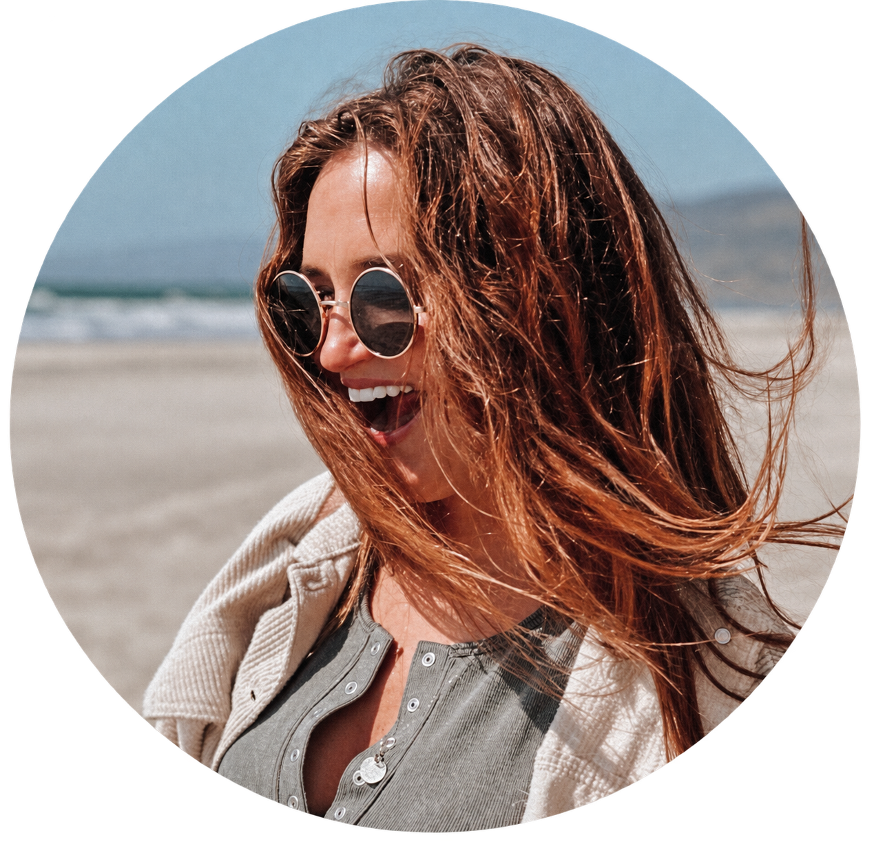 woman in sunglasses smiles while wind blows her hair on a california beach