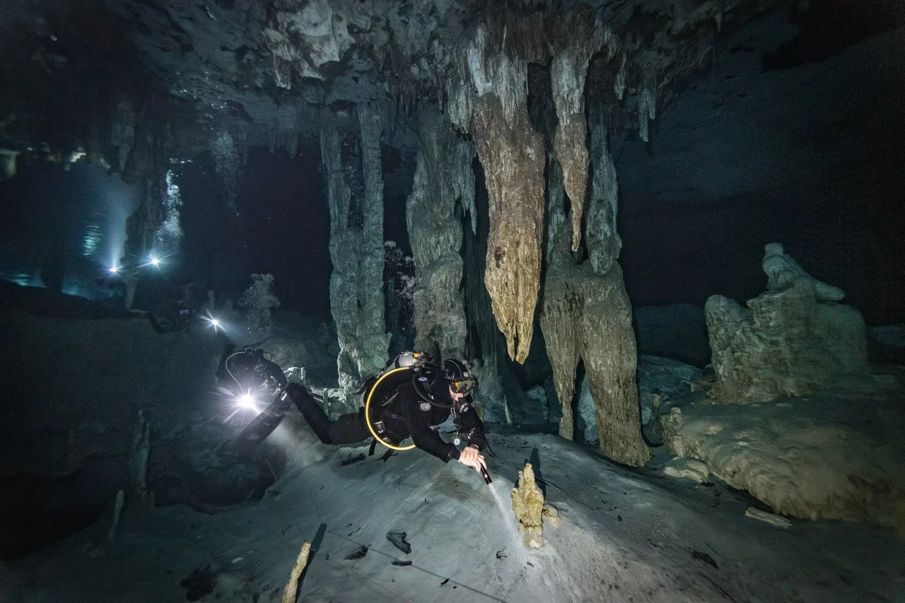 A scuba diver is exploring inside a dark underground cave with stalactites hanging from the ceiling and stalagmites rising from the floor, illuminated by their underwater lights.