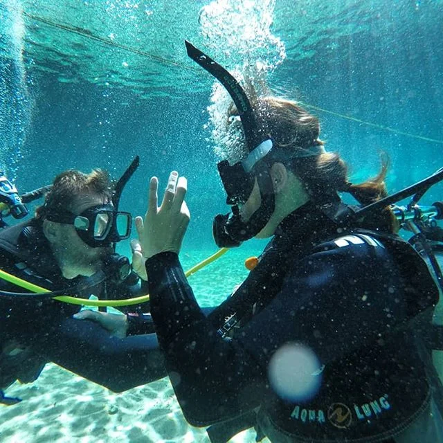 Two people scuba diving underwater, facing each other, with one giving a peace sign and both wearing masks and wetsuits.