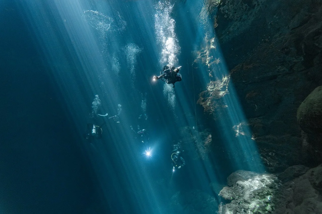 scuba divers descend through beams of light in el pit cenote