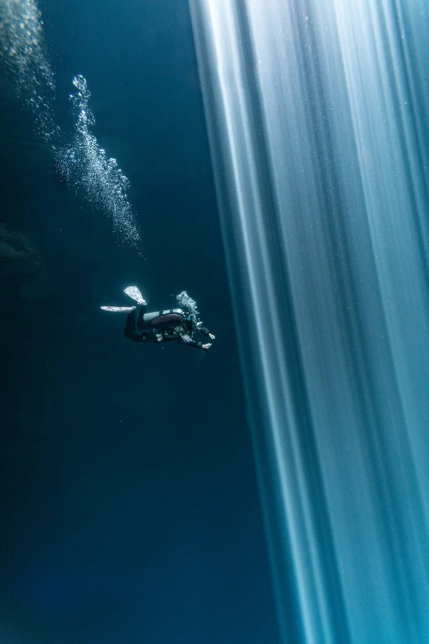 Scuba diver swimming in an underwater cave or crevice, with sunlight streaming through from above.
