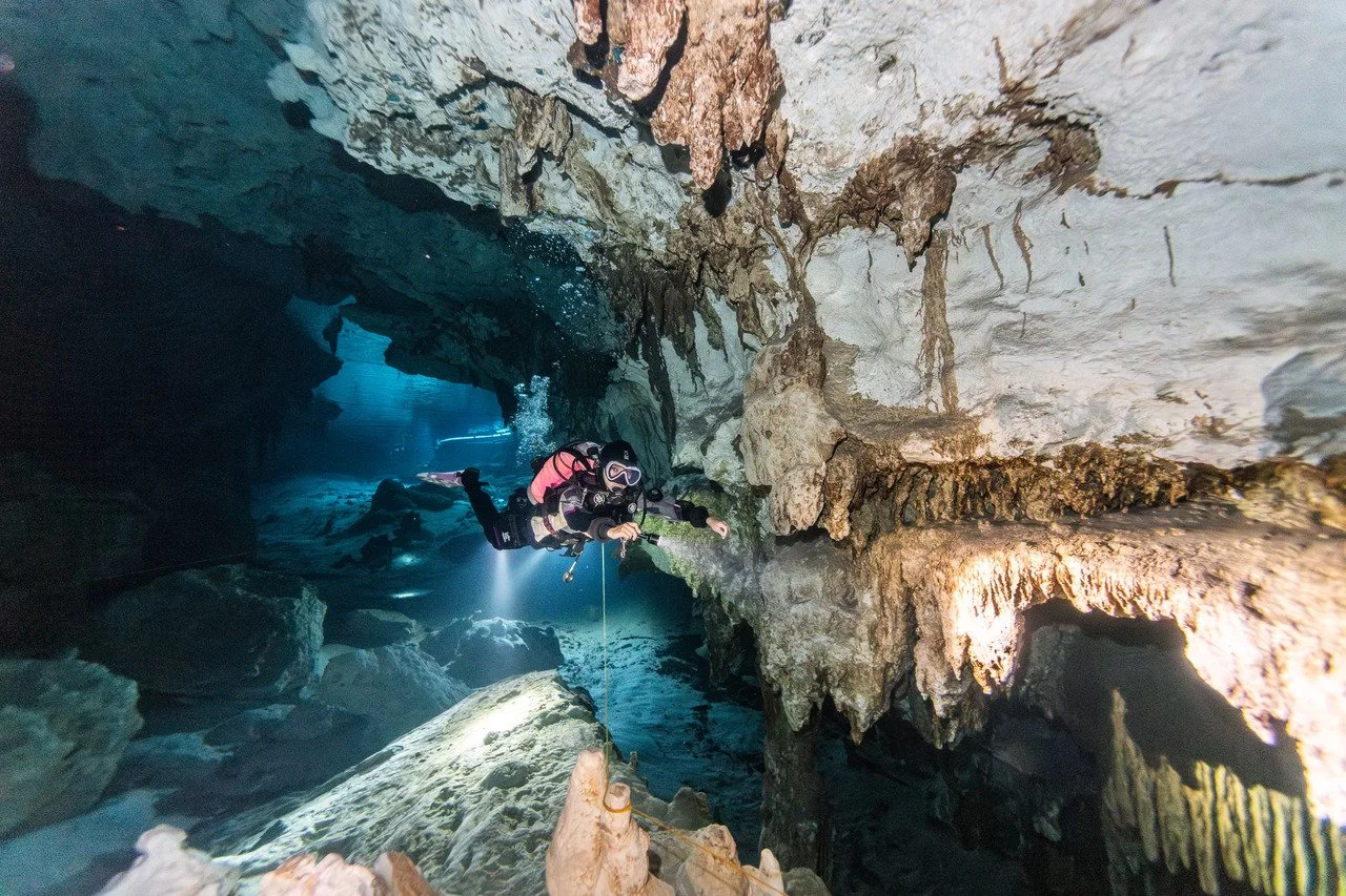 A scuba diver exploring an underwater cave filled with rocks and stalactites, illuminated by artificial lights.