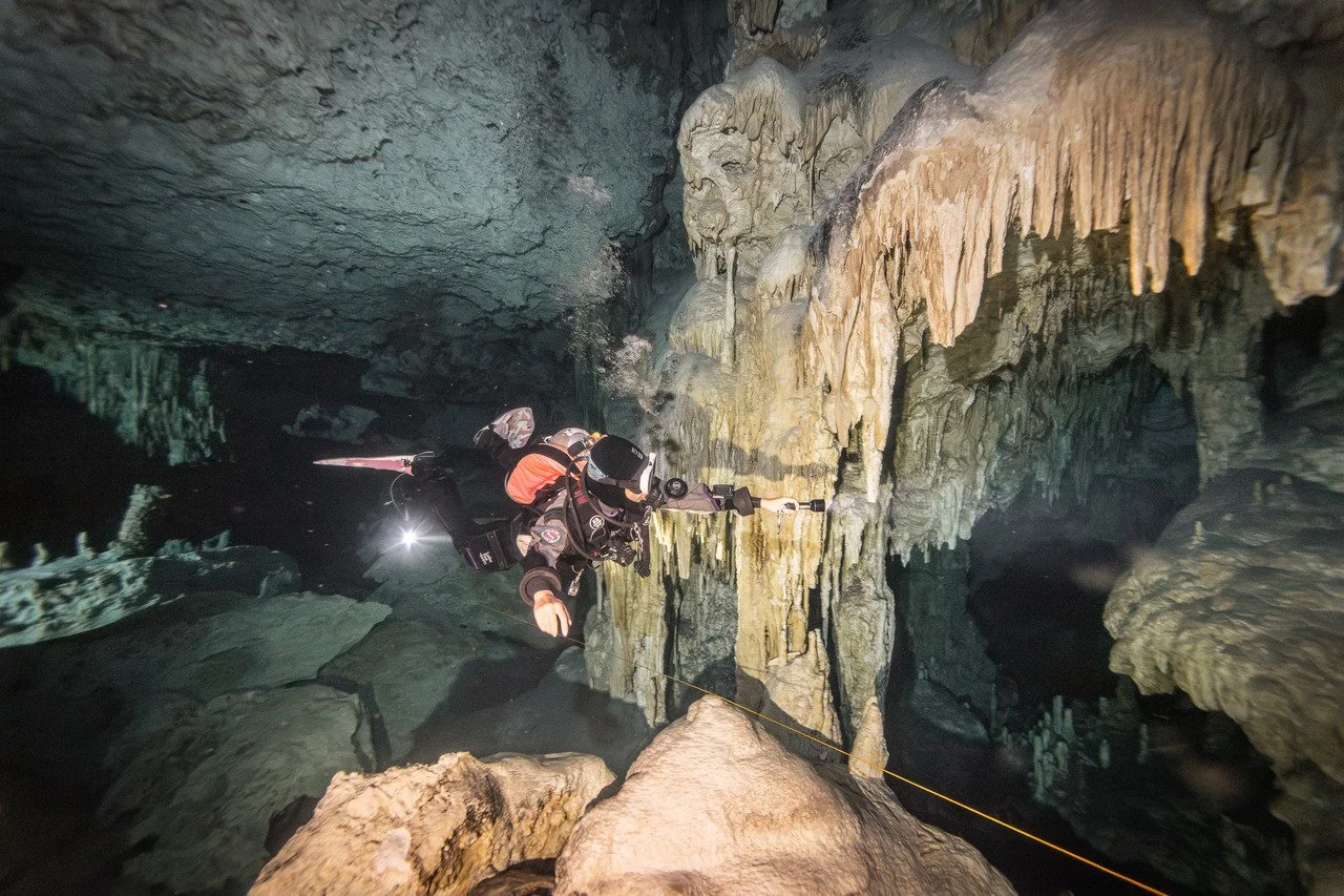 A person in caving gear exploring an underground cavern with stalactites and stalagmites.