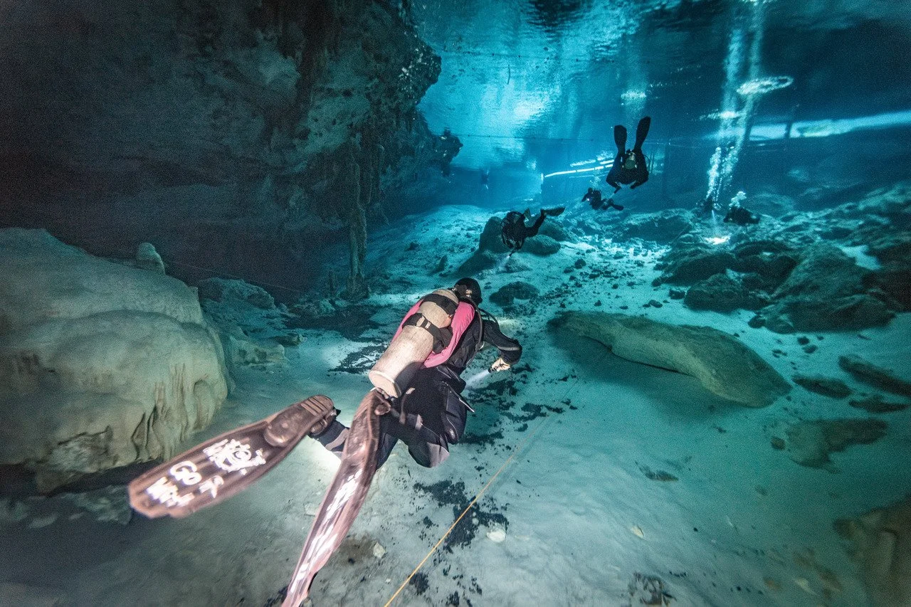 Scuba divers exploring an underwater cave with rocky walls and water-lit ceiling.