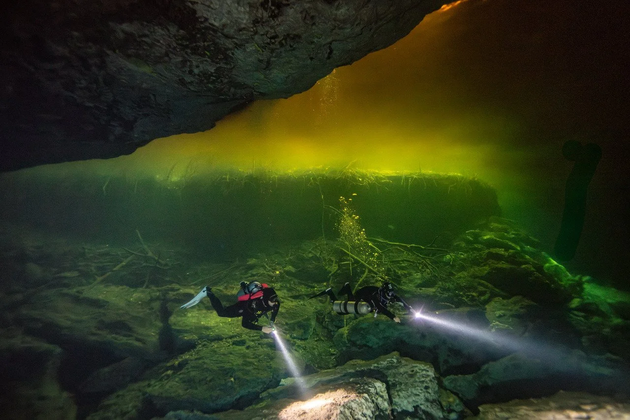 Two divers explore an underwater cave with a yellowish glow from an opening above, using flashlights to illuminate the rocky floor and debris.