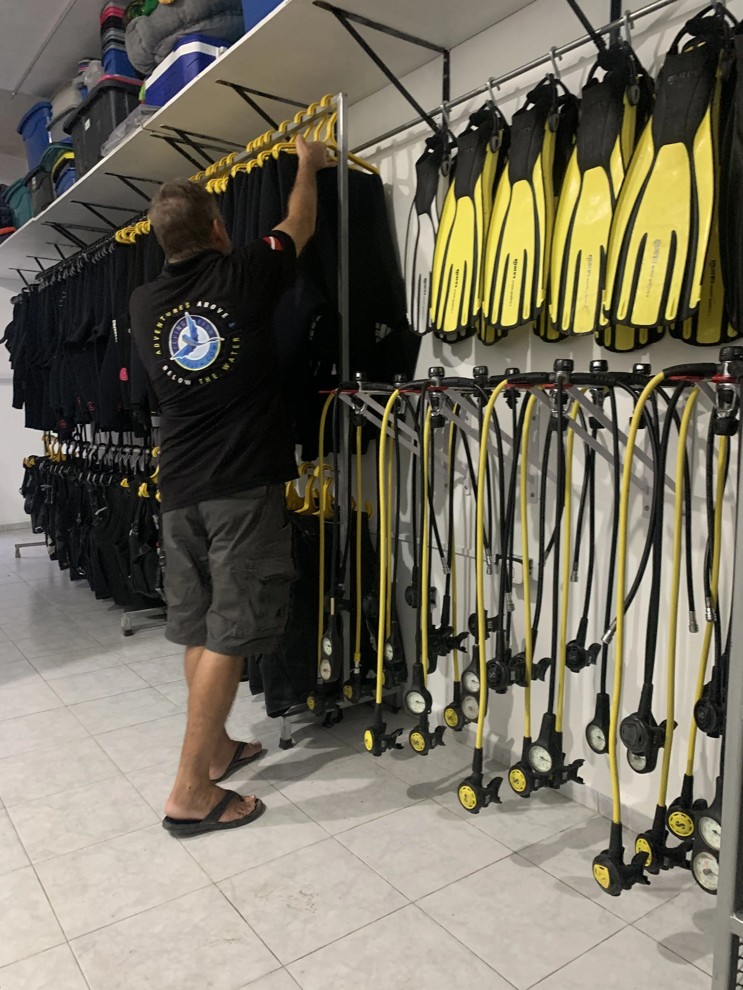 A person browsing outdoor gear in a store, standing barefoot in flip-flops, wearing a black shirt with a logo on the back. The store has shelves with bags and a wall with fire hoses or similar equipment.