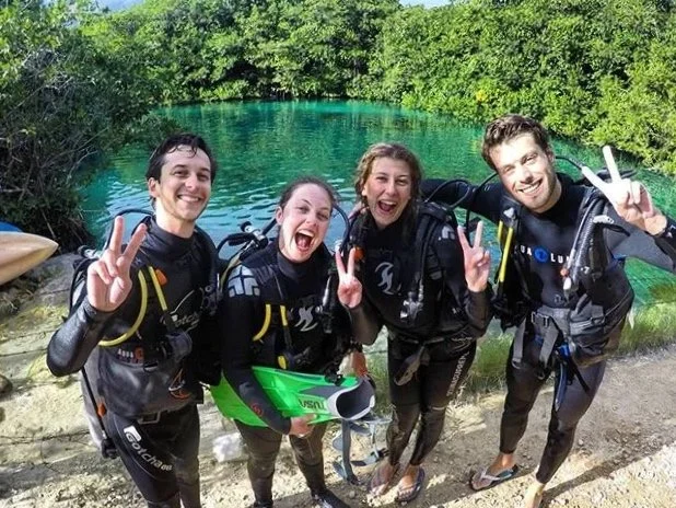 Three scuba divers exploring underwater cave with rocks and aquatic plants.