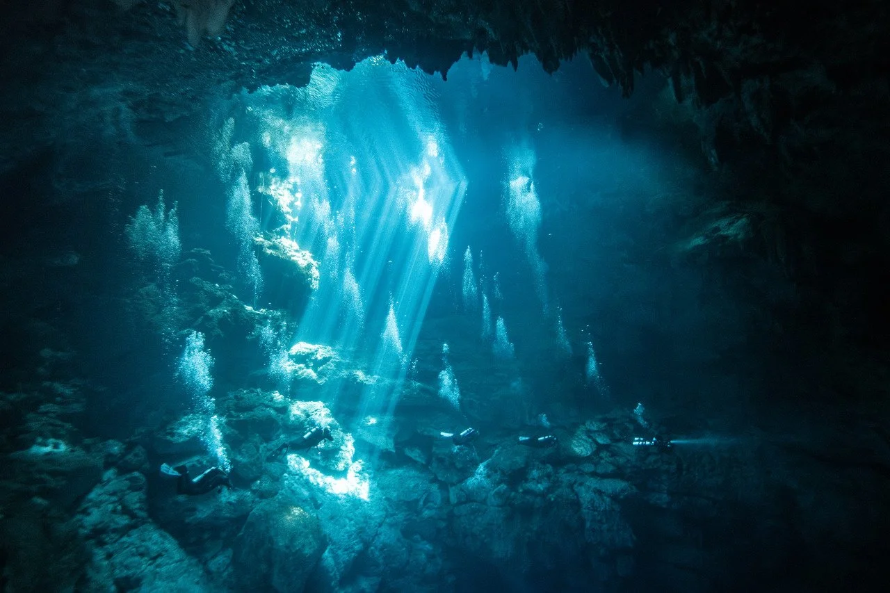 Underwater scene with sunlight streaming through cave opening, revealing divers exploring the rocky cave interior.