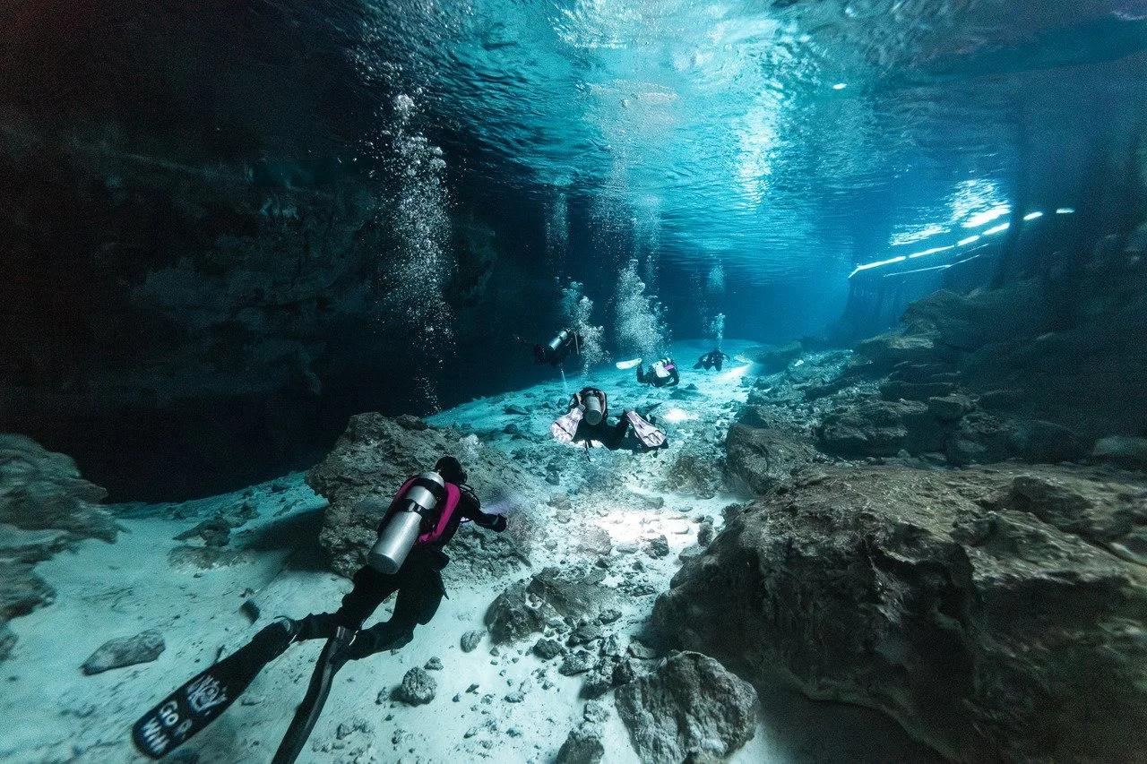 Divers exploring an underwater cave or cavern with rocky surroundings and clear blue water.