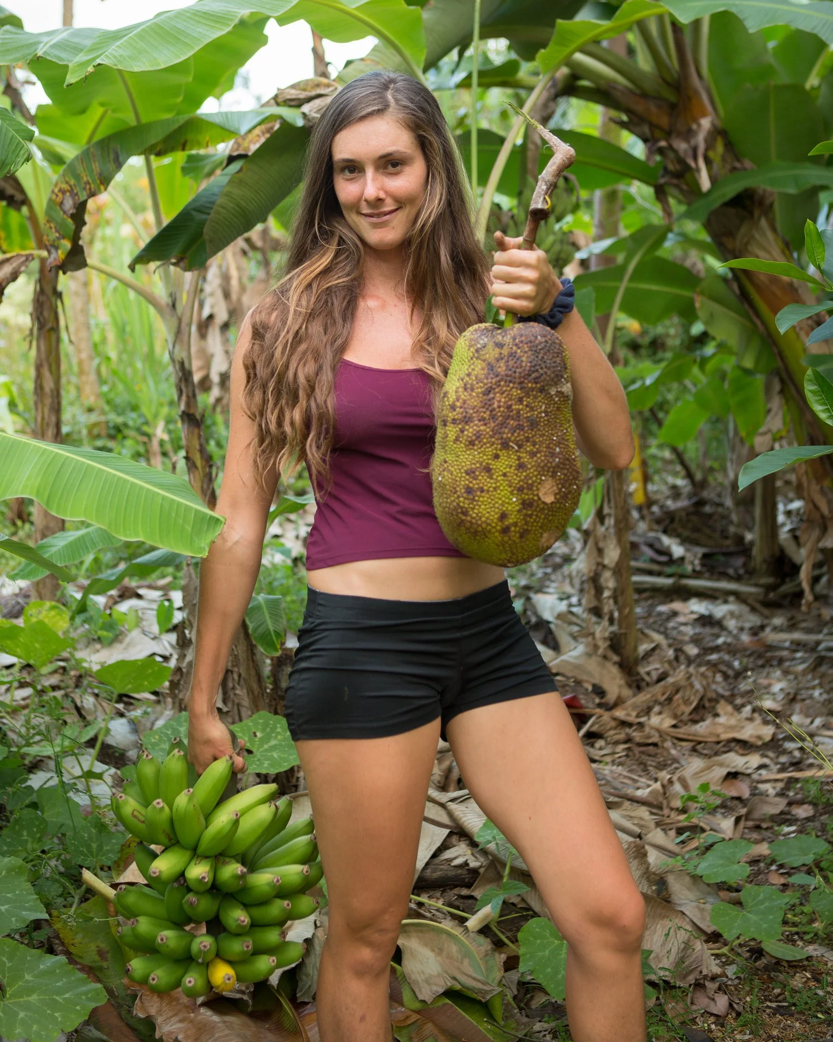 A woman standing in a banana plantation holding a large jackfruit in her right hand and a bunch of green bananas in her left hand.