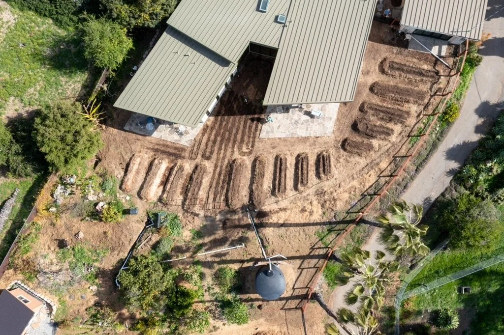 Bird's eye view of a backyard with dirt planting beds, a covered patio with outdoor furniture, and a fenced perimeter with greenery.