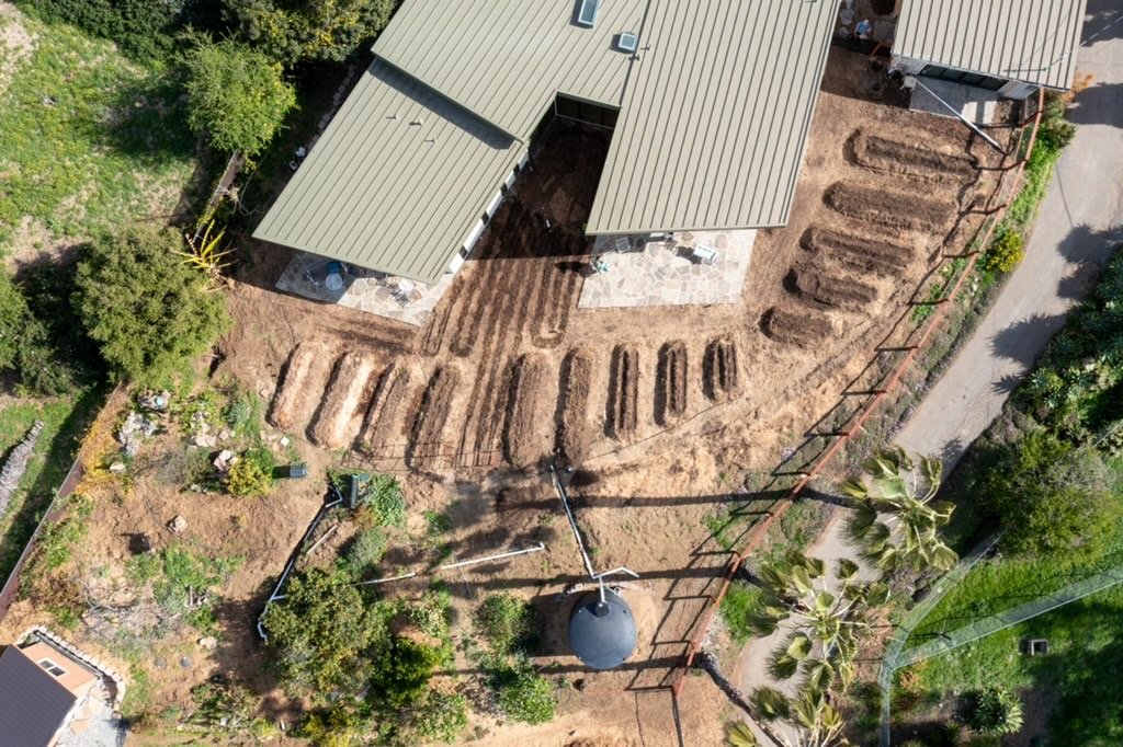 Aerial view of a backyard with landscaping in progress, garden beds, trees, and a house with a metal roof.