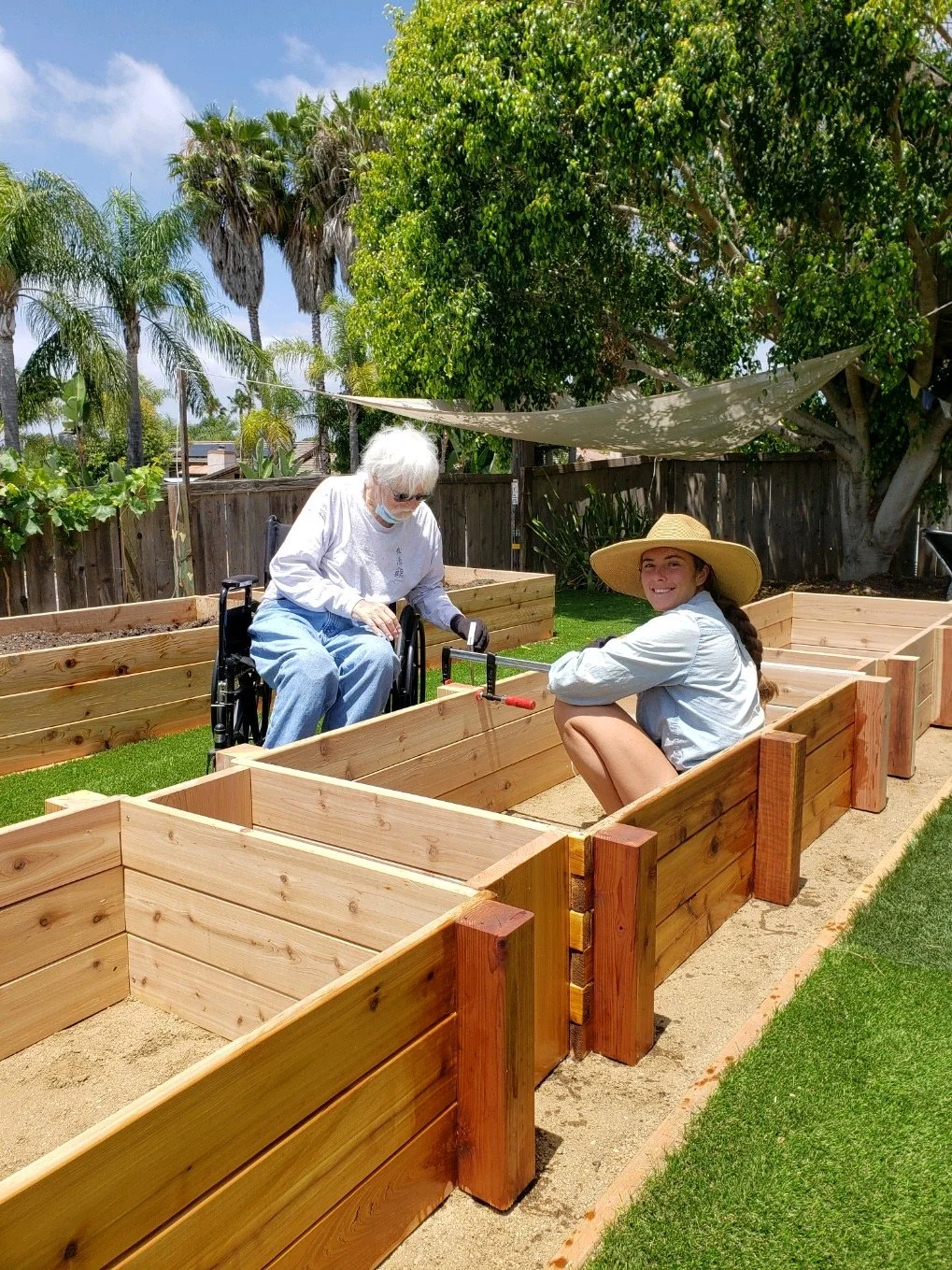 Two women working in a garden with wooden planters, one seated in a wheelchair and the other squatting, wearing a wide-brimmed hat. Trees and a fence are in the background.