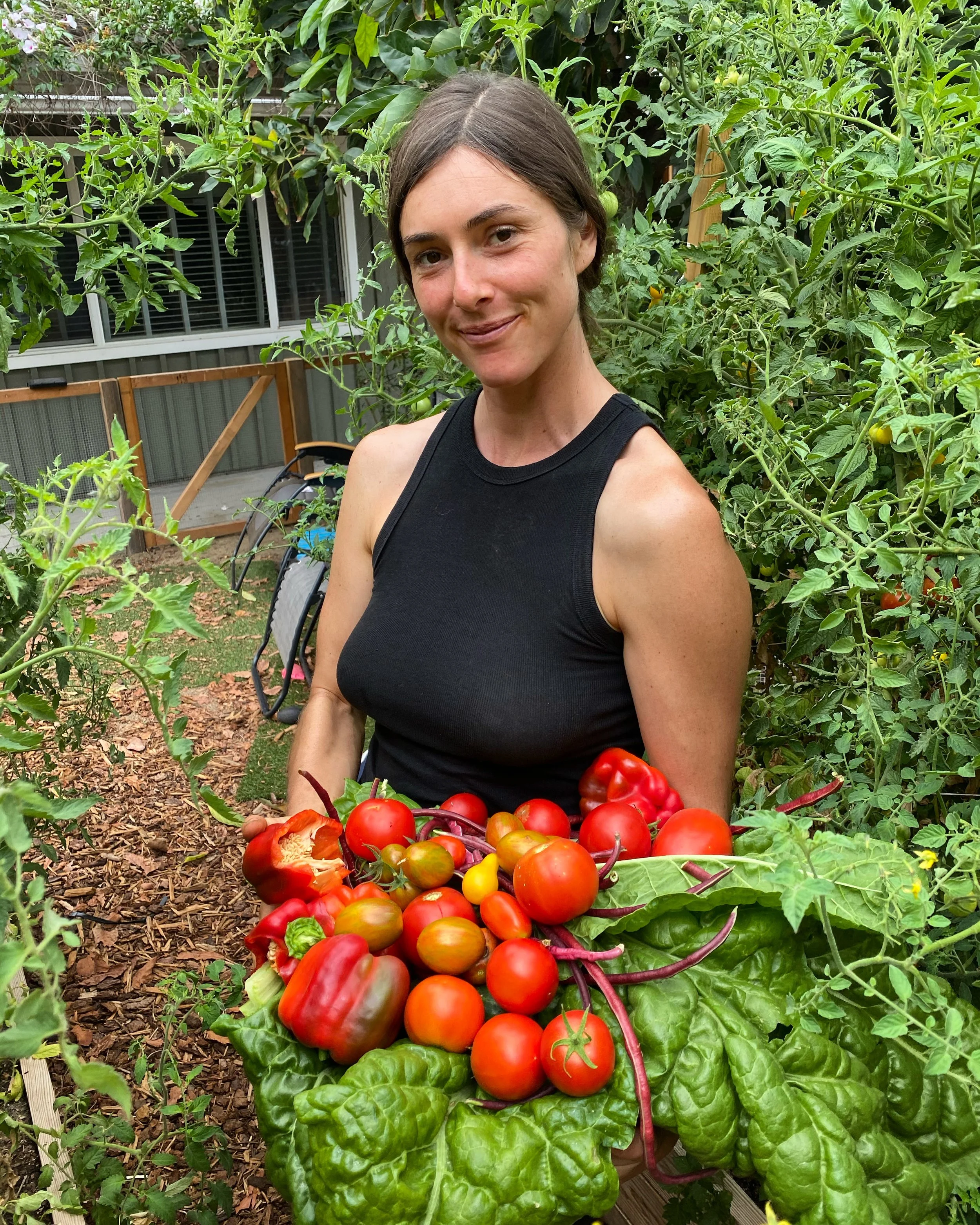A woman standing in a garden holding a large bunch of freshly picked tomatoes and leafy greens.
