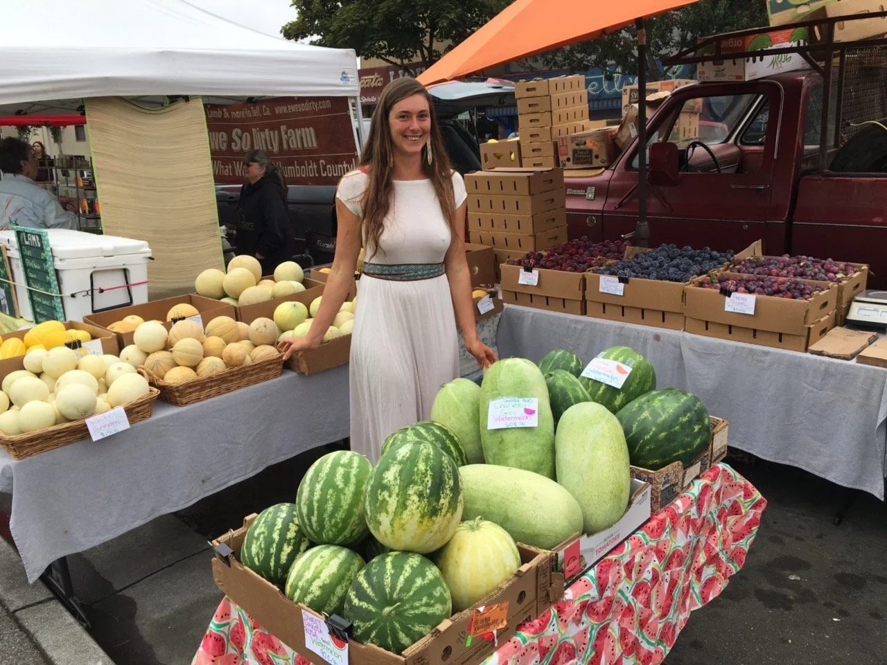 A woman in a white dress smiling at a farmers market standing next to a display of large watermelons in a cardboard box. Behind her, there are various fruits including melons, grapes, and cardboard boxes with produce. Market tents and other vendors are visible in the background.