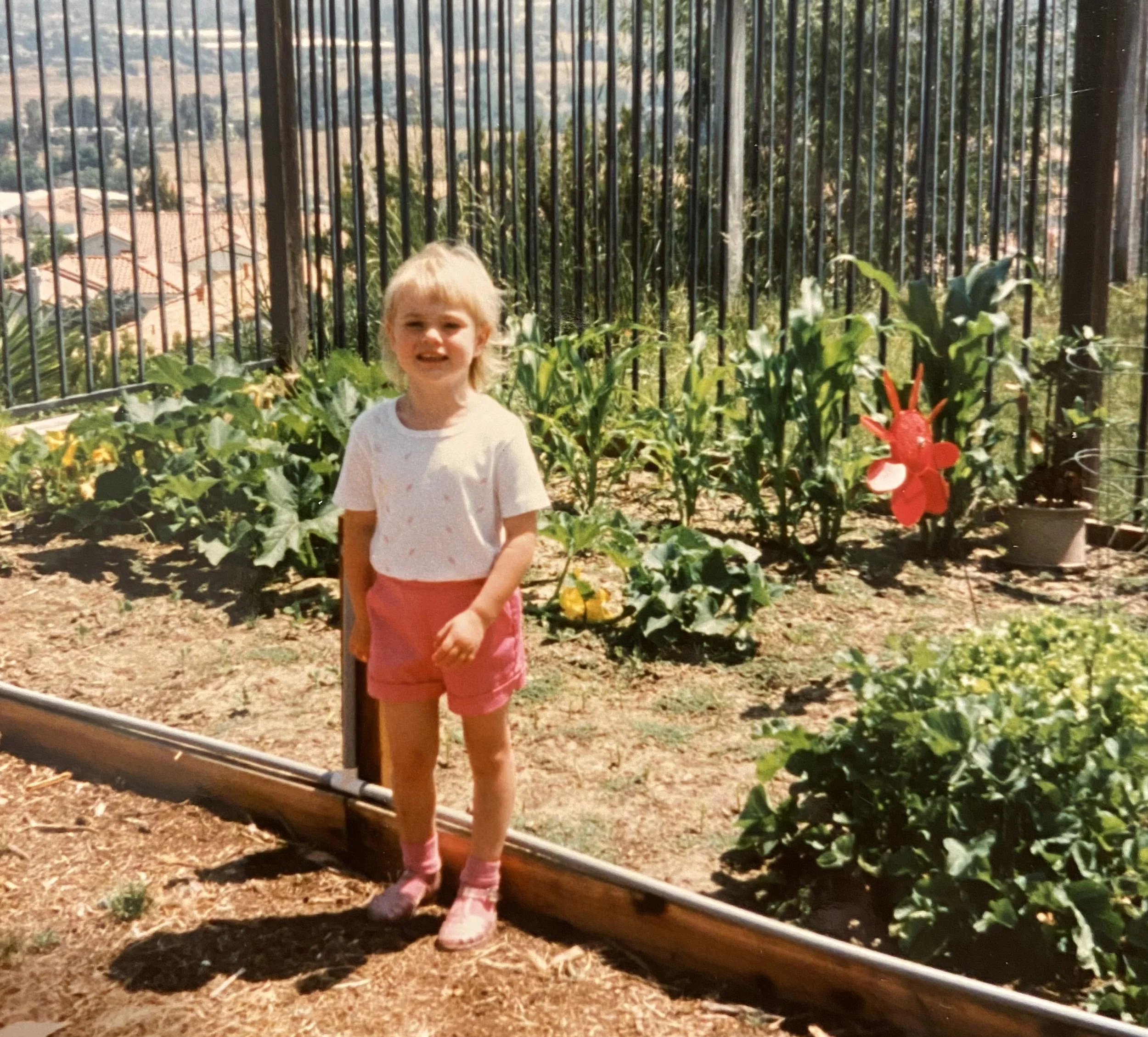A young girl with blonde hair standing on a garden path, smiling, surrounded by flowers and greenery, with a metal fence in the background.