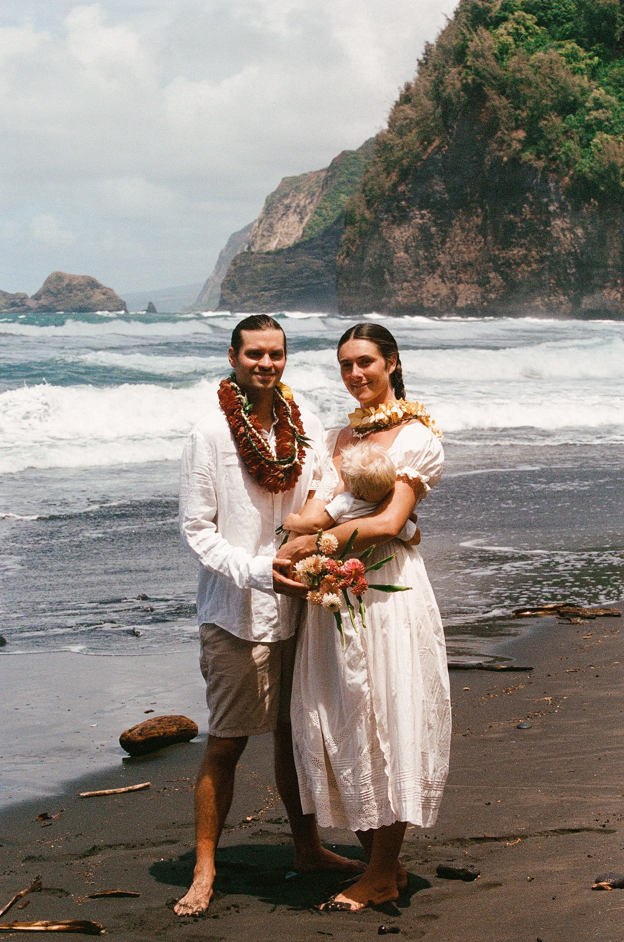 A couple in wedding attire standing on a black sand beach, holding a child, with large green cliffs and ocean waves in the background, traditional leis around their necks.