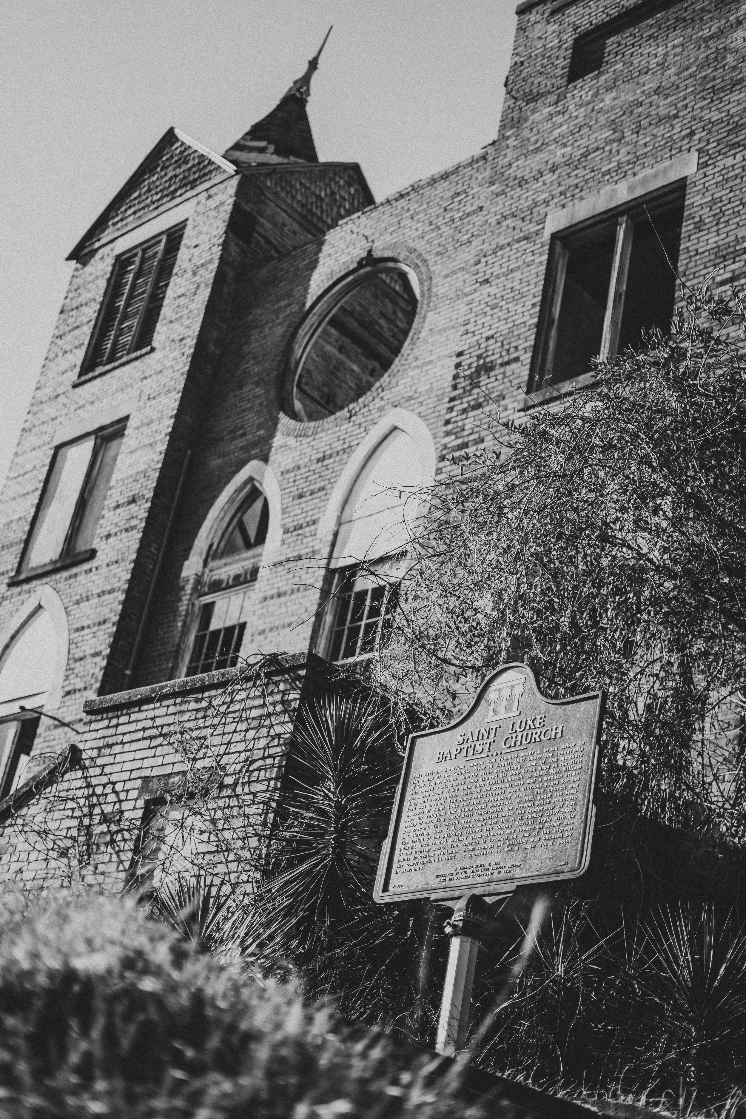 Black and white photo of Saint Luke Baptist Church, a brick building with arched windows, a tall steeple, and a visible sign providing historical information about the church.