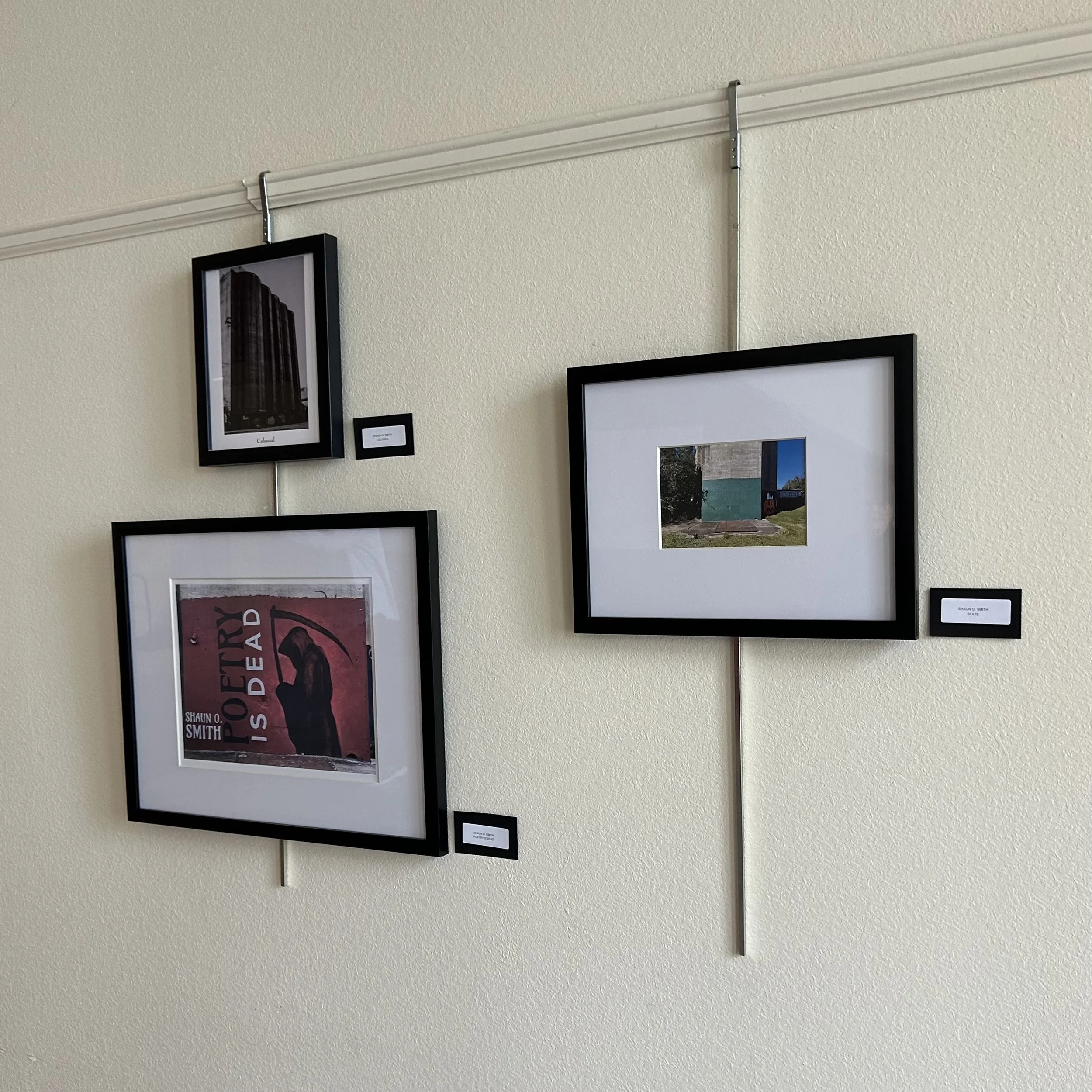 Multiple framed photographs and artworks are displayed on a beige wall, arranged in a grid pattern.