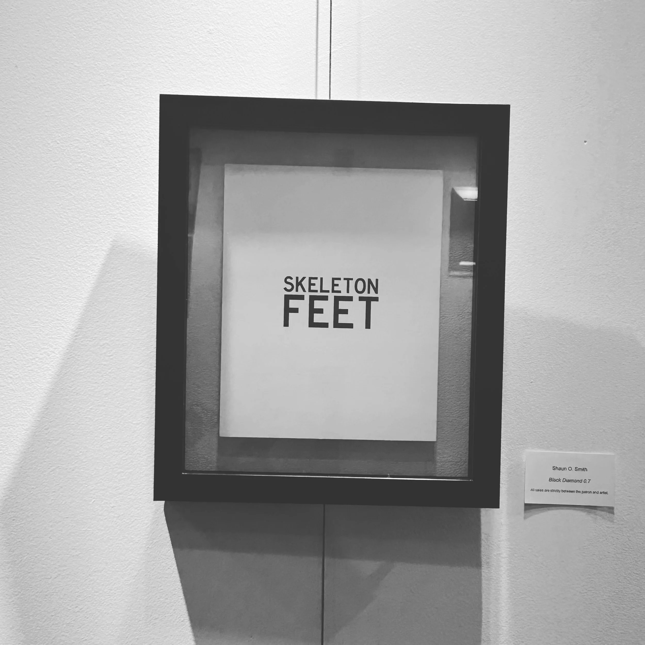 Front of a white poster with bold black text that reads "SKELETON FEET." The poster is attached to a wall, and there is a small printed label with text in the bottom right corner. The wall is painted in a light color, and the poster appears to be par