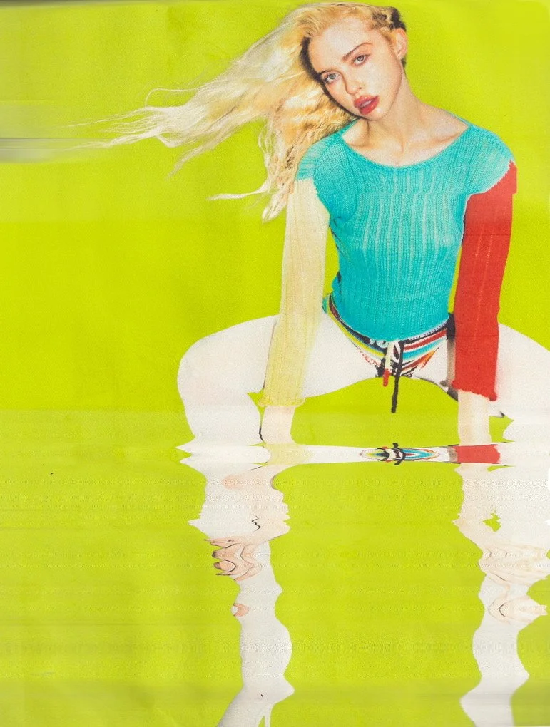 A woman with long blond hair, wearing a colorful sweater and striped shorts, sitting on a reflective surface with a bright green background.