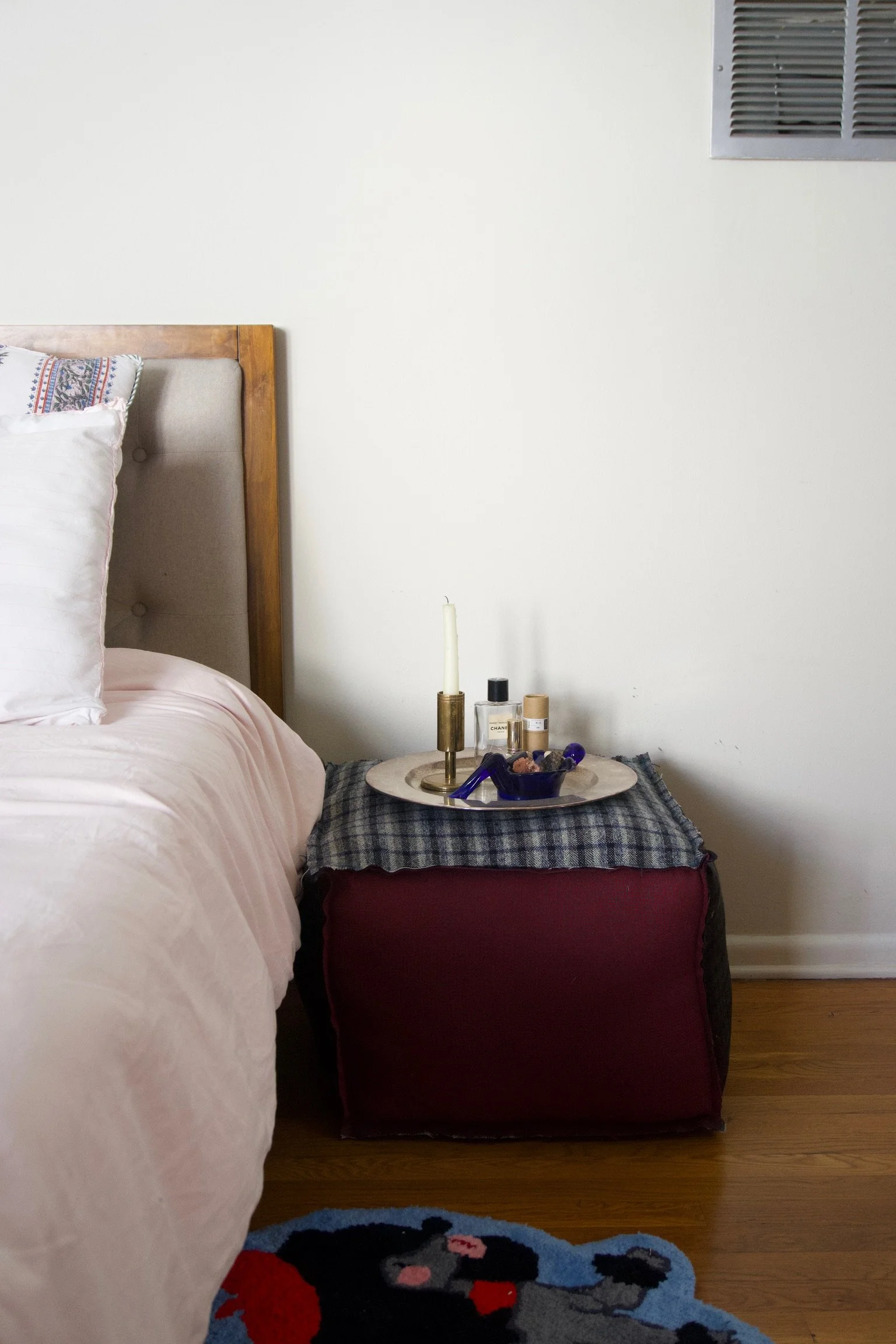 Close-up of a beige bed with a pink duvet and white pillows beside a red fabric storage cube with a checkered cloth on top. A round tray with perfume, a candle, and a candle holder with a candle is on the cloth. Part of a patterned rug is visible on 