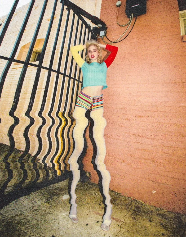 A woman standing against a brick wall, wearing colorful striped shorts, a light blue cropped sweater, and long white thigh-high socks. The photo has a distorted, wavy effect.