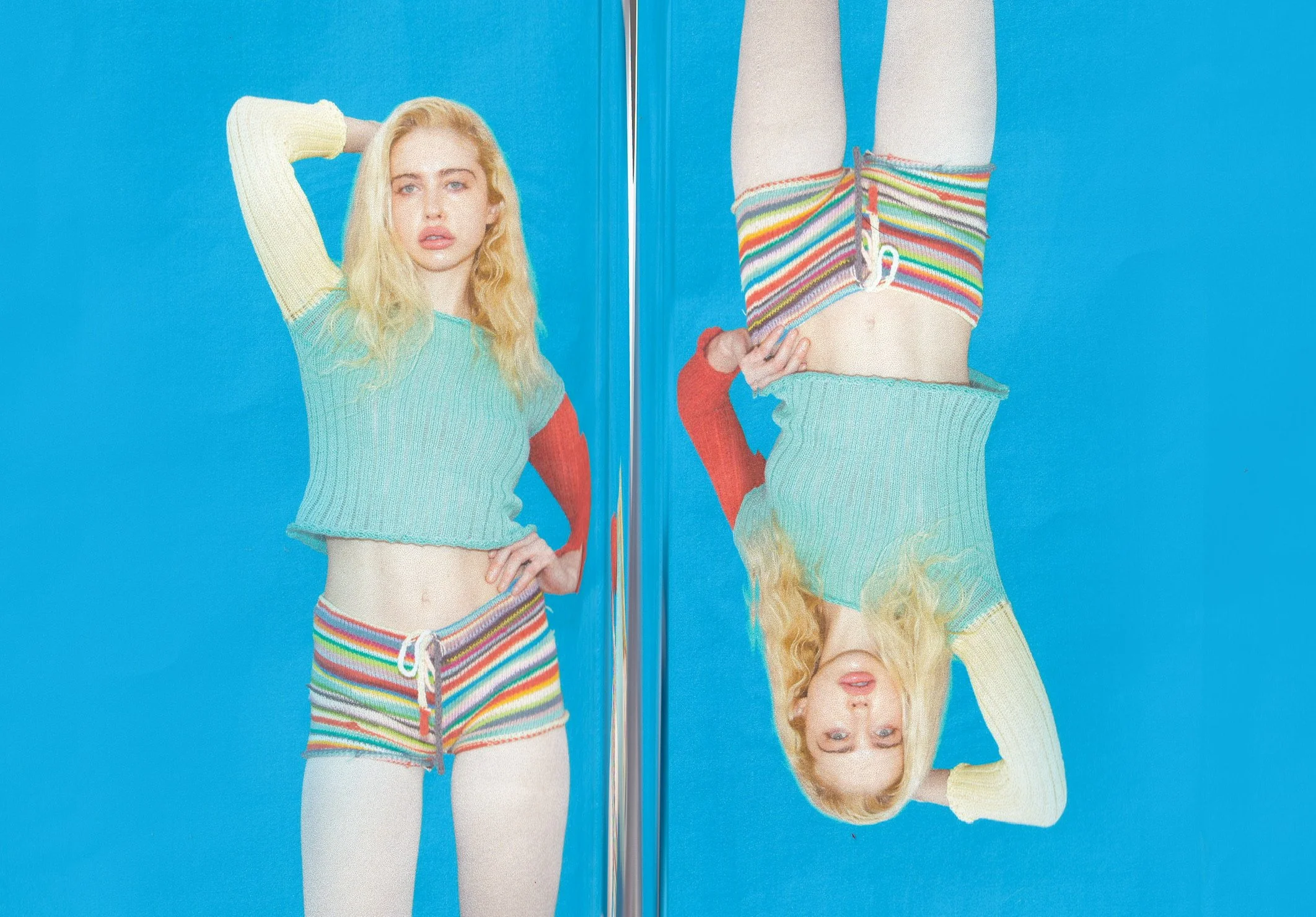 A young woman with blonde hair posing against a bright blue background. She is wearing a multicolored striped pair of shorts, a multicolored striped crop top, white tights, and a long-sleeve knit sweater with red sleeves, with her left hand on her hi