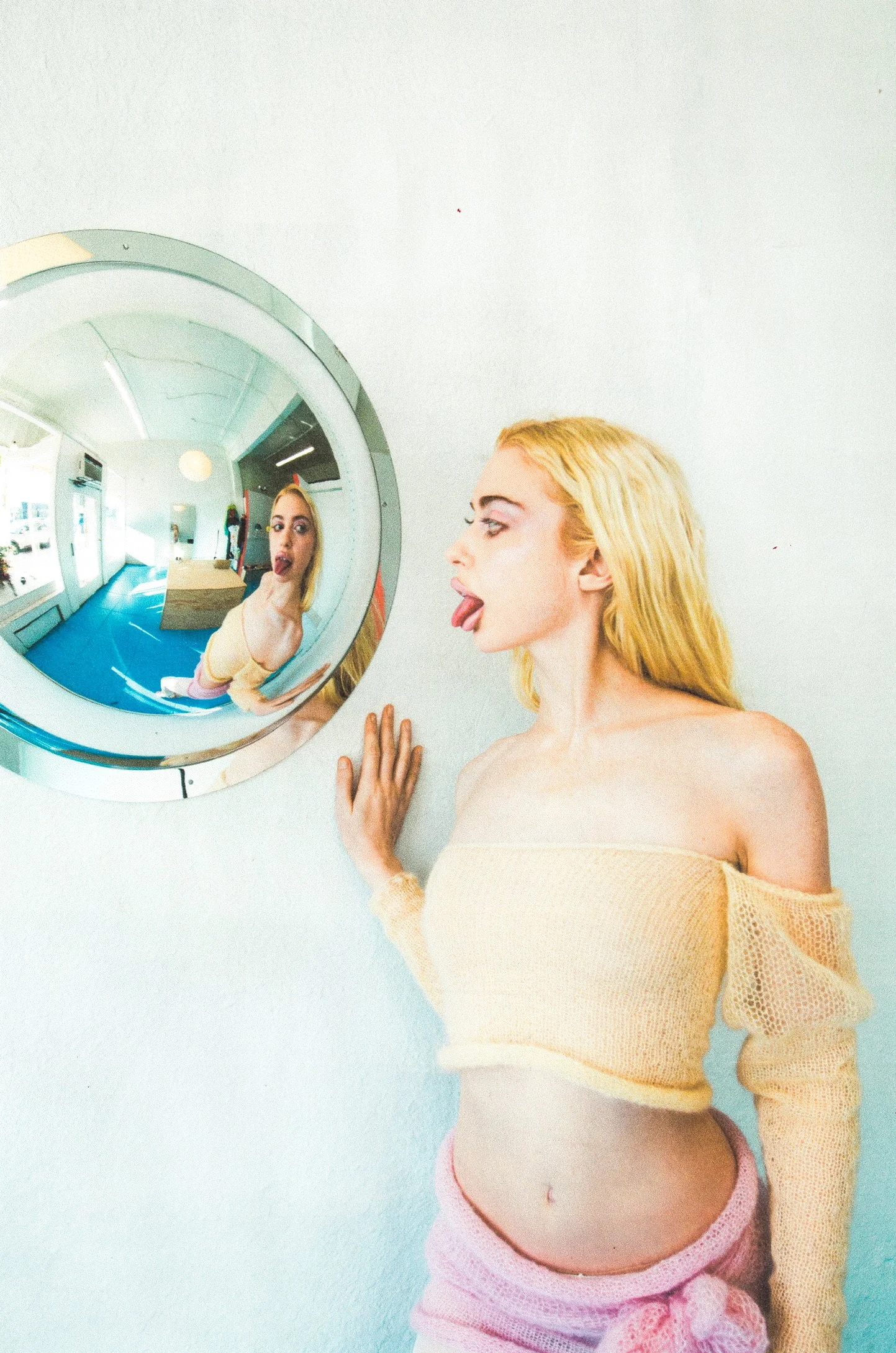A woman with long blonde hair sticking her tongue out while looking at her reflection in a round mirror on a white wall.