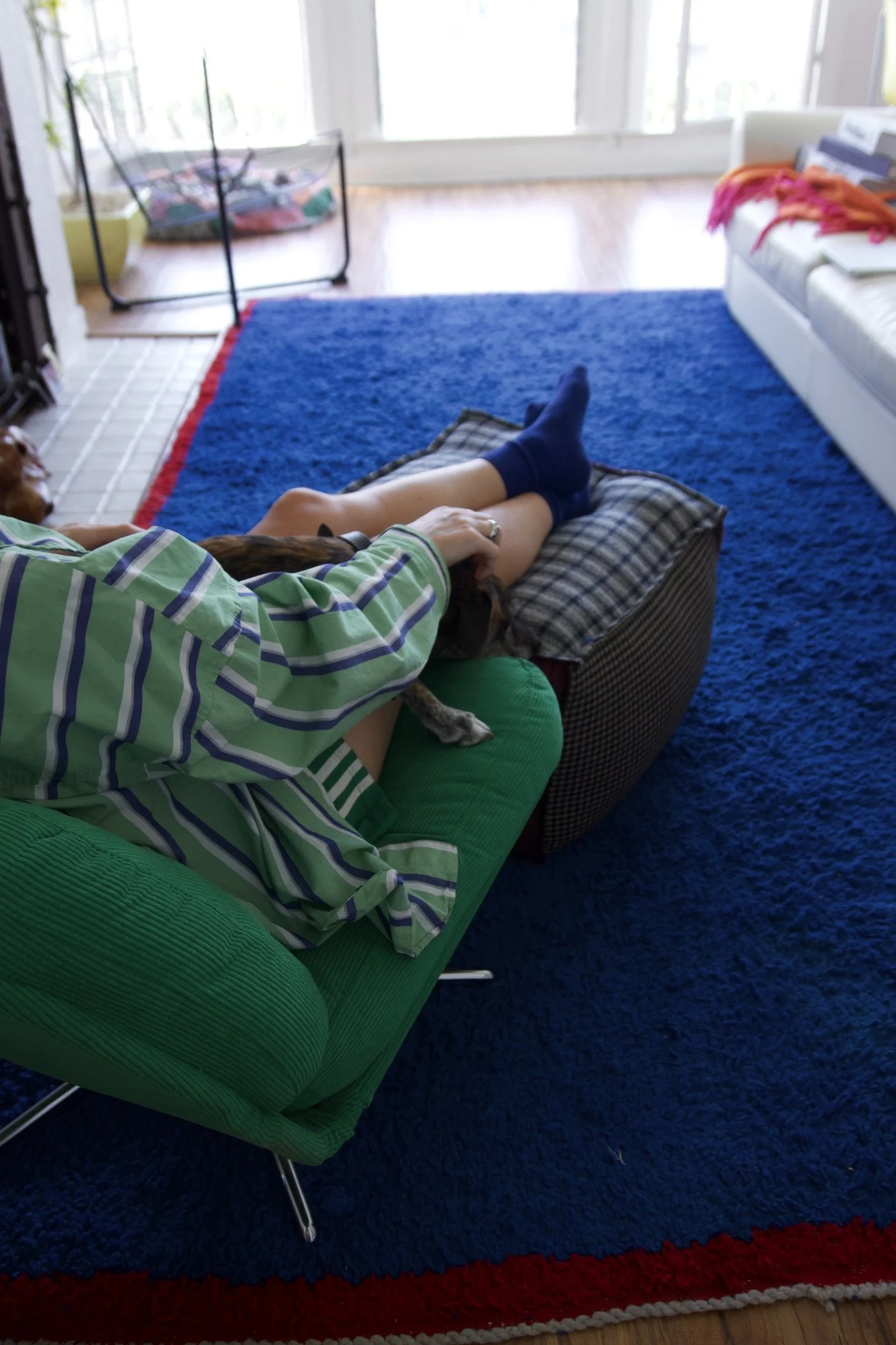 Person lying on a green couch with legs outstretched, petting a dog on a cushion in a living room with a blue rug, white sofa with clothes on it, and a sunny balcony in the background.