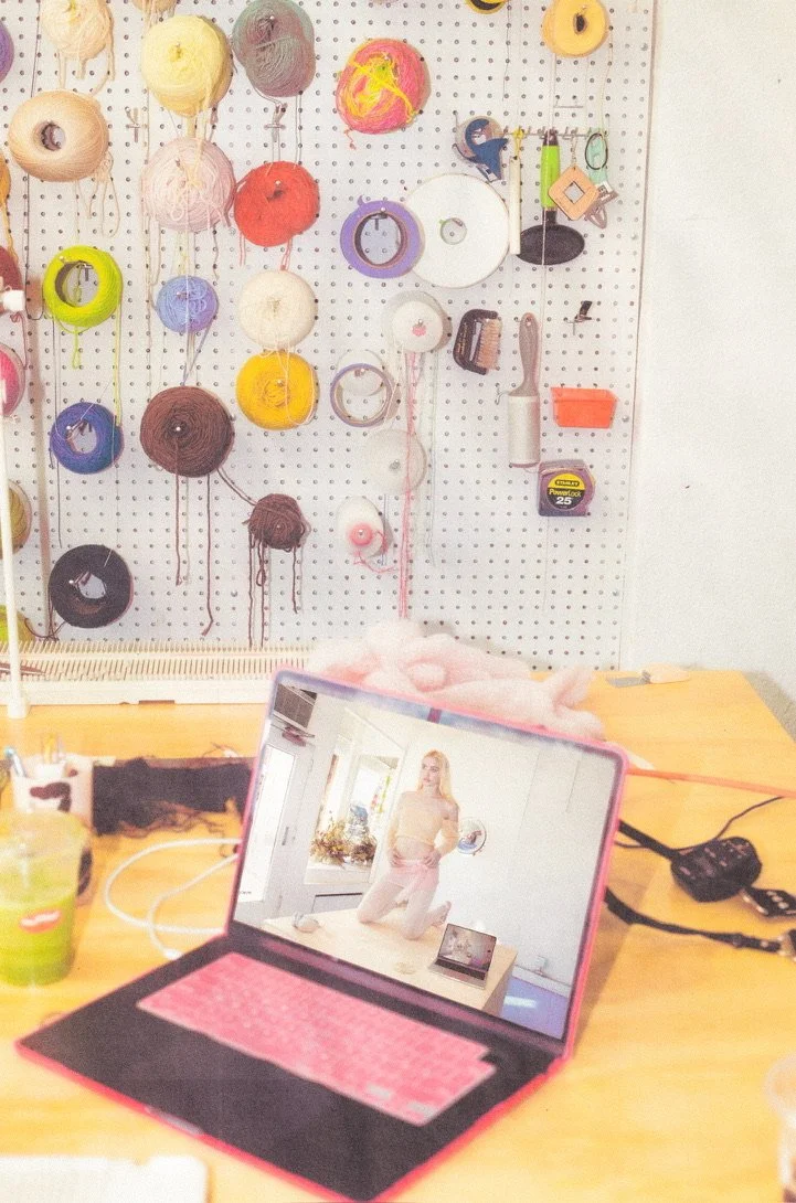 A workspace with a computer showing a woman in a beige sweater and pants kneeling on a table, with colorful yarn spools and tools hanging on a pegboard in the background.