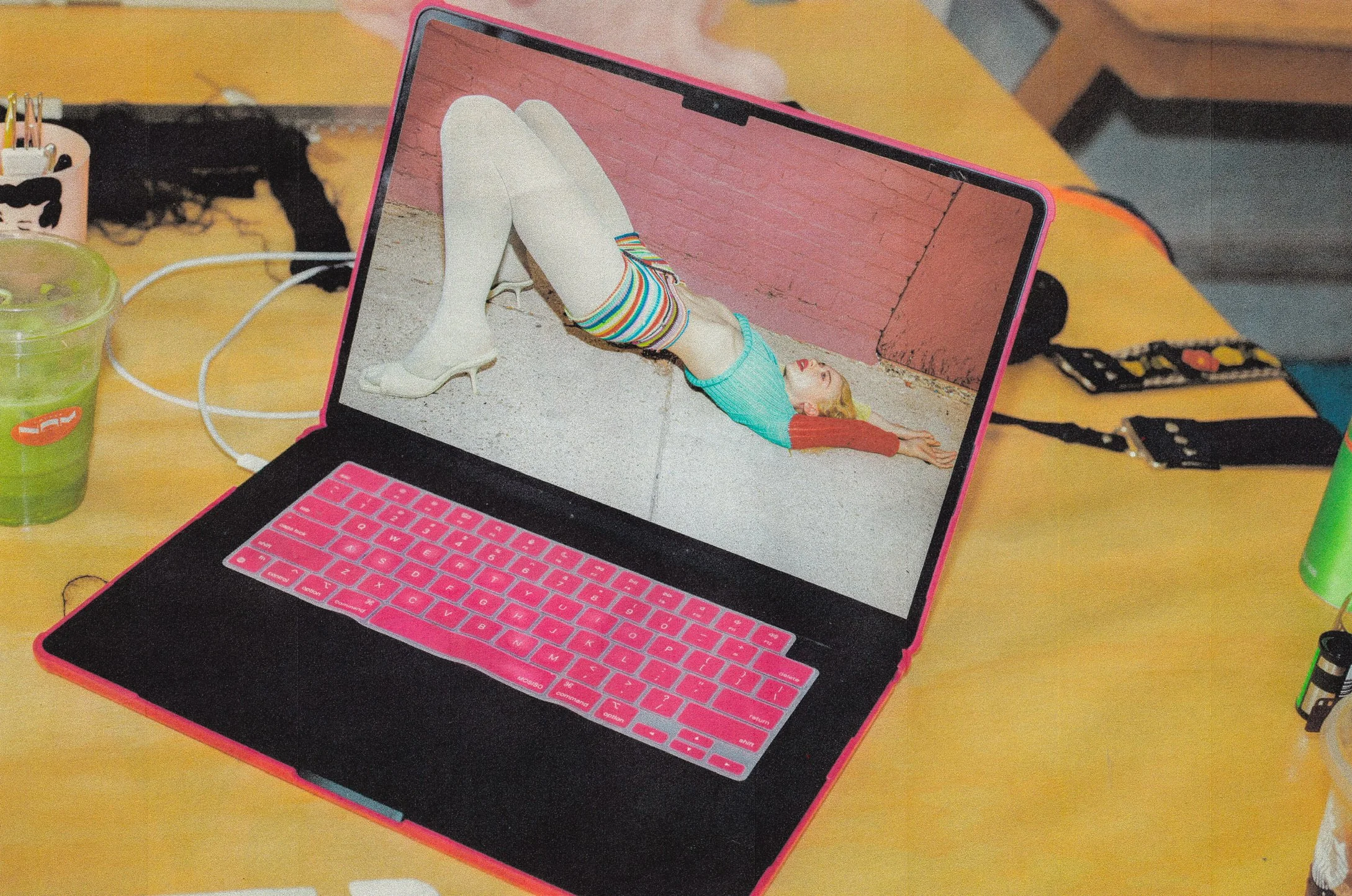 Laptop with a pink keyboard cover on a wooden desk displaying a woman lying on the ground in colorful clothing and striped tights, against a pink brick wall background.