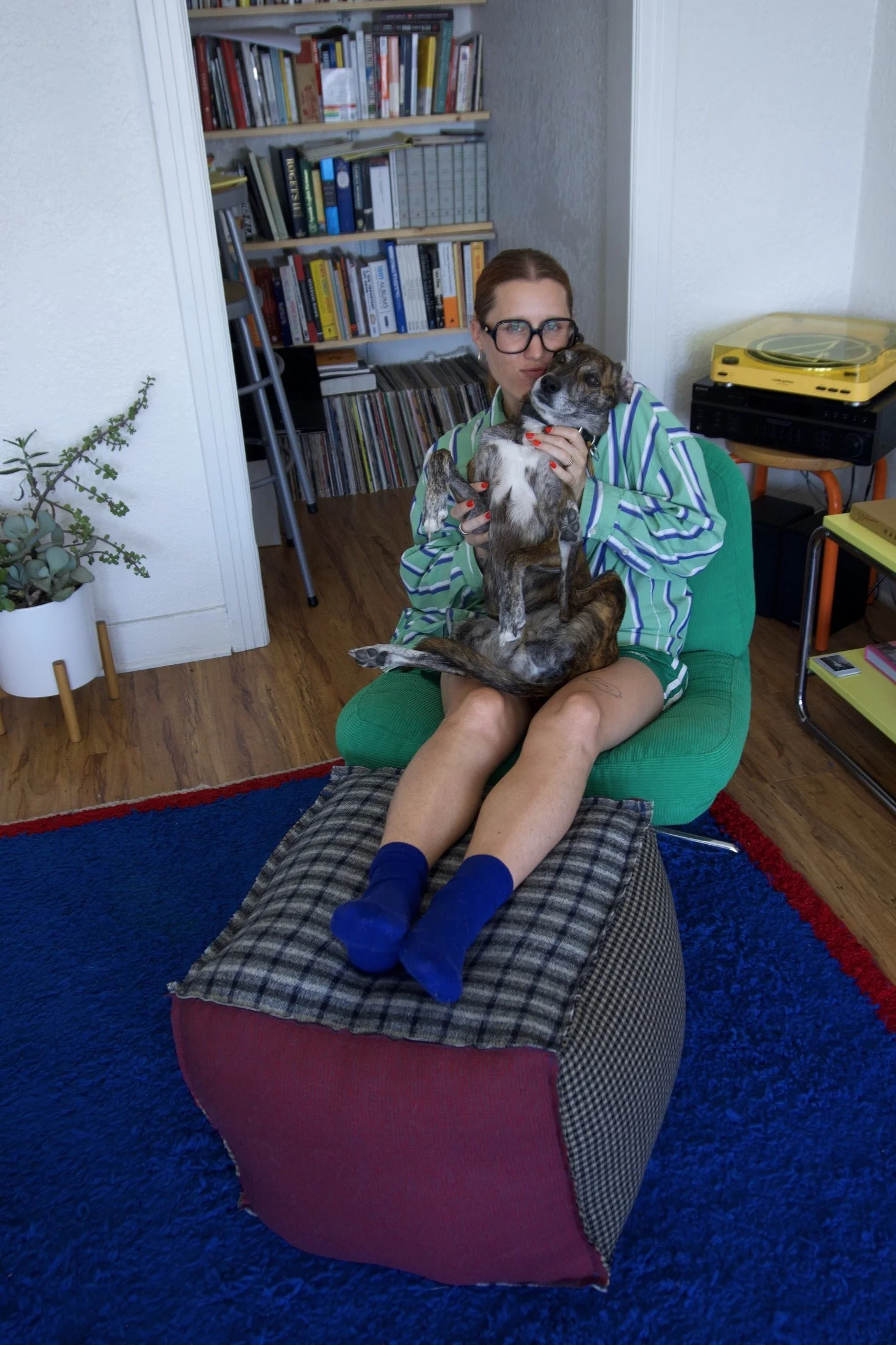 A woman with glasses and red nail polish sitting on a green chair, holding a small dog in her lap. The woman is wearing a striped shirt, and there is a bookshelf filled with books and vinyl records behind her. She is sitting on a strange large, multi