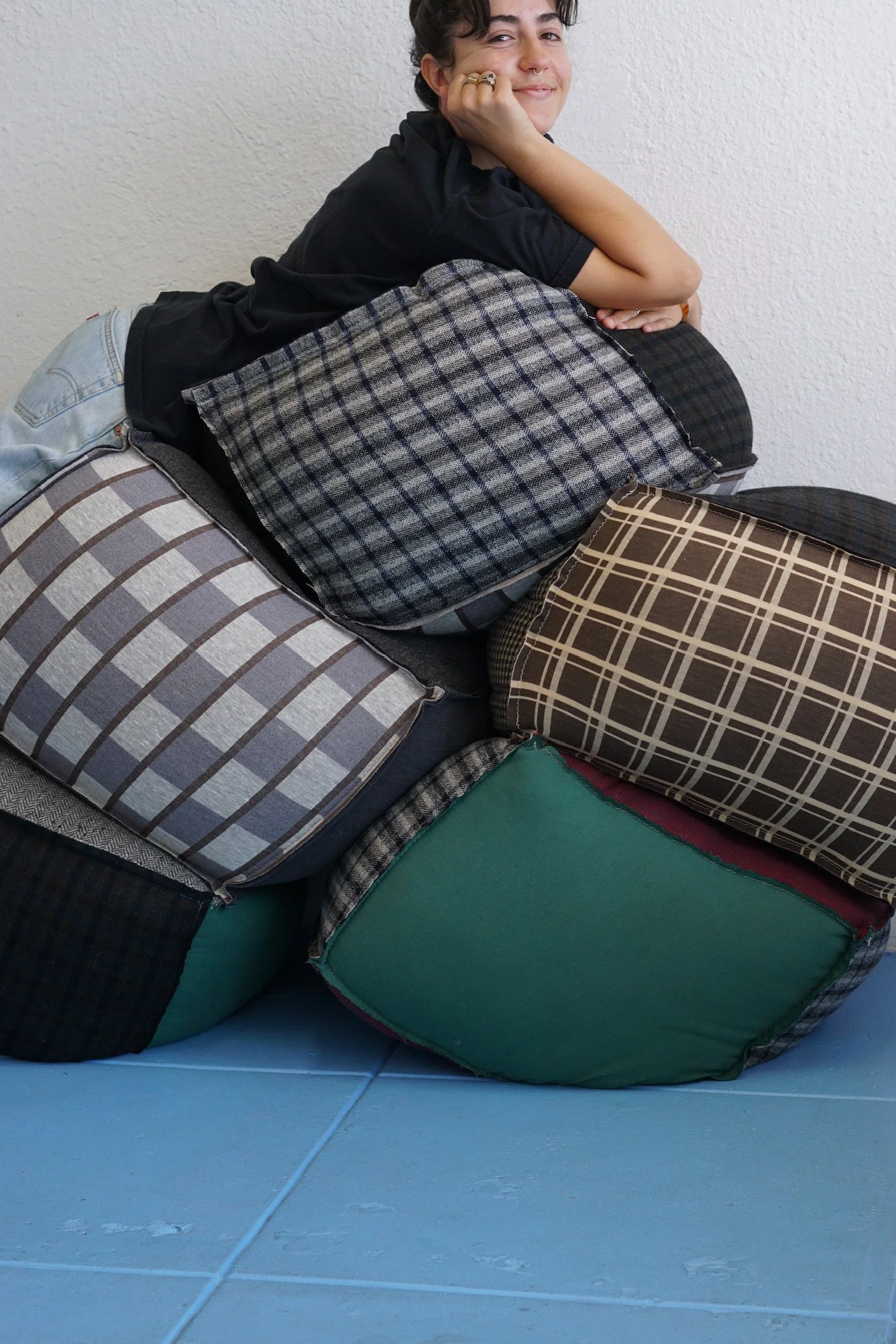 Person resting their head on their hand, smiling, while lying on a stack of various patterned cushions and pillows against a white wall.